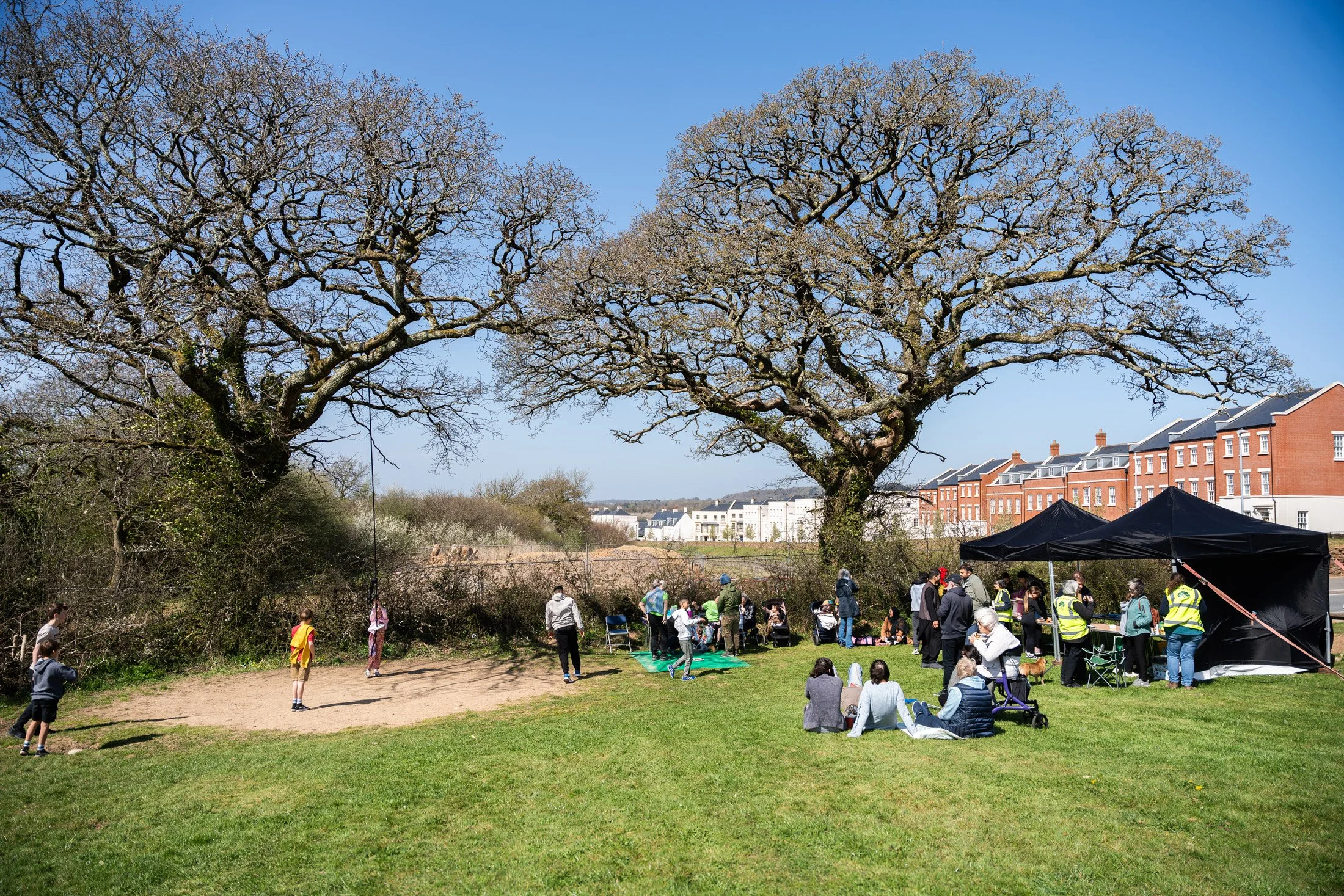 Children and adults playing and gathering in a park on a sunny day, with tall leafless trees and residential buildings in the background.
