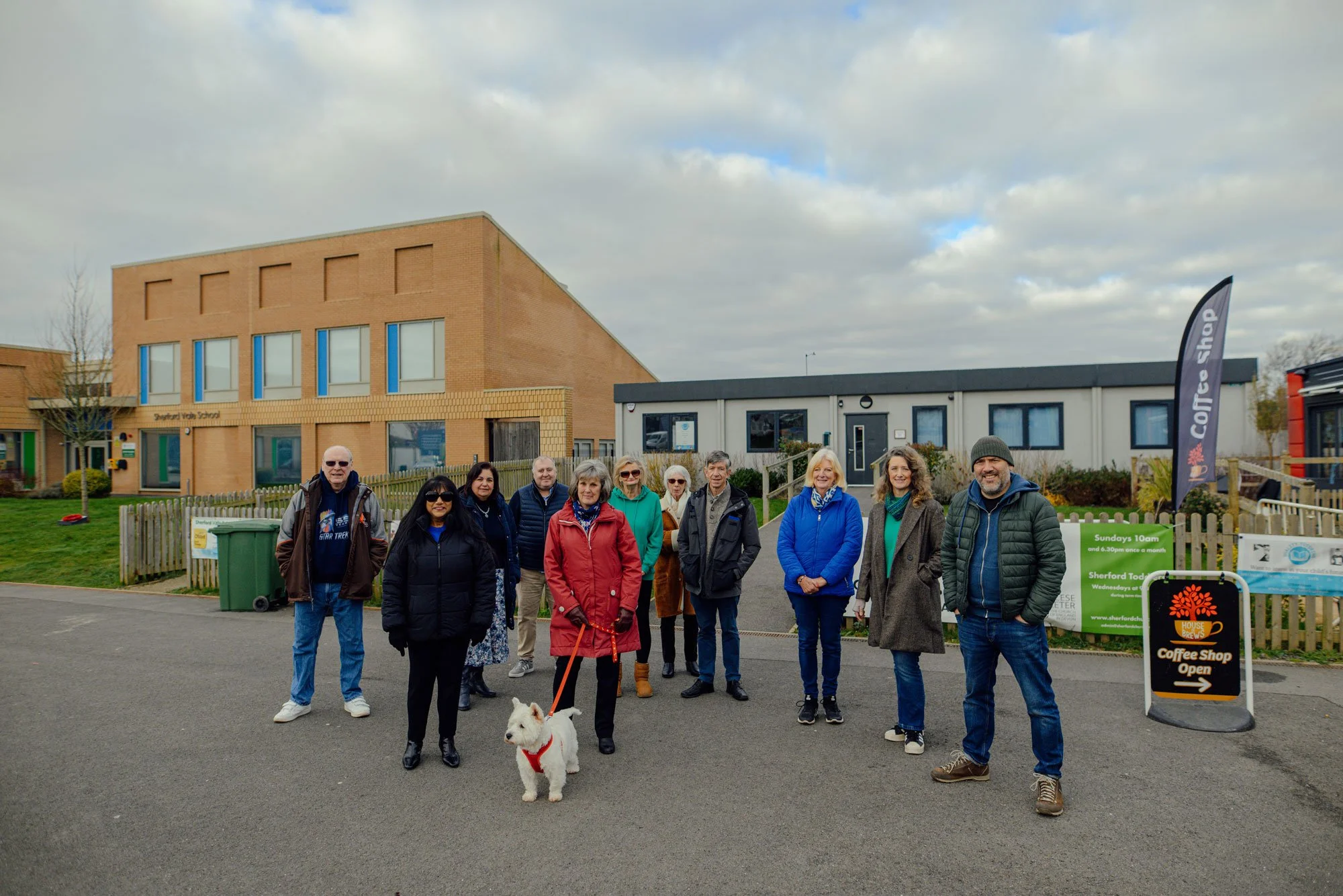 A group of people standing outdoors in front of a building with signs indicating a coffee shop and a school, with a dog on a leash in the foreground.
