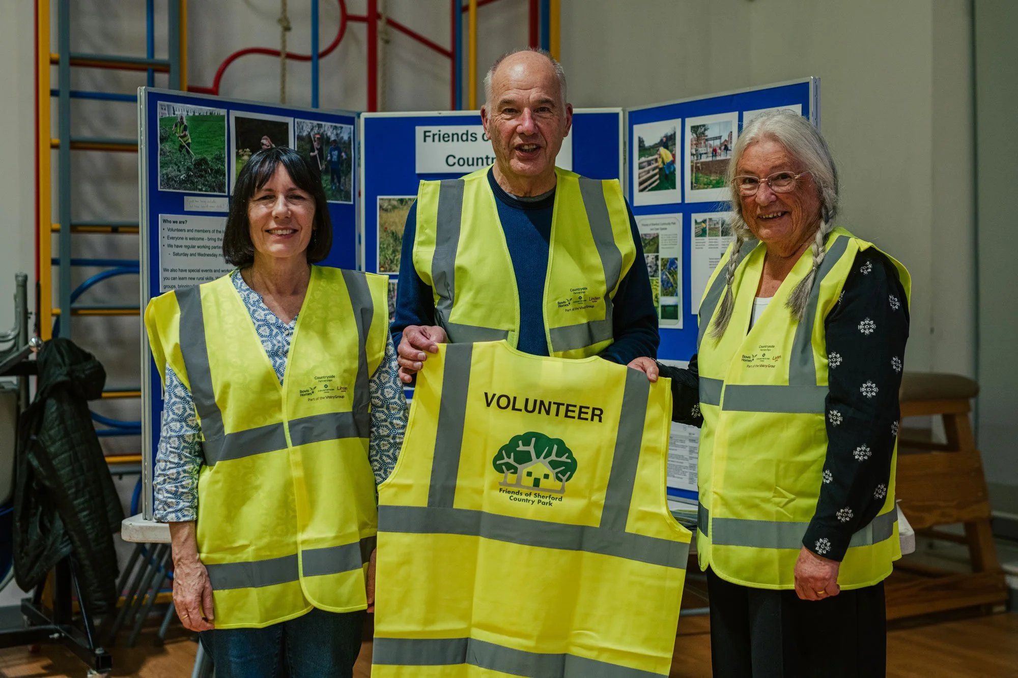 Three women and one man wearing yellow volunteer vests standing indoors in front of a display board, holding a yellow volunteer vest with the logo of Friends of Sherford Country Park.