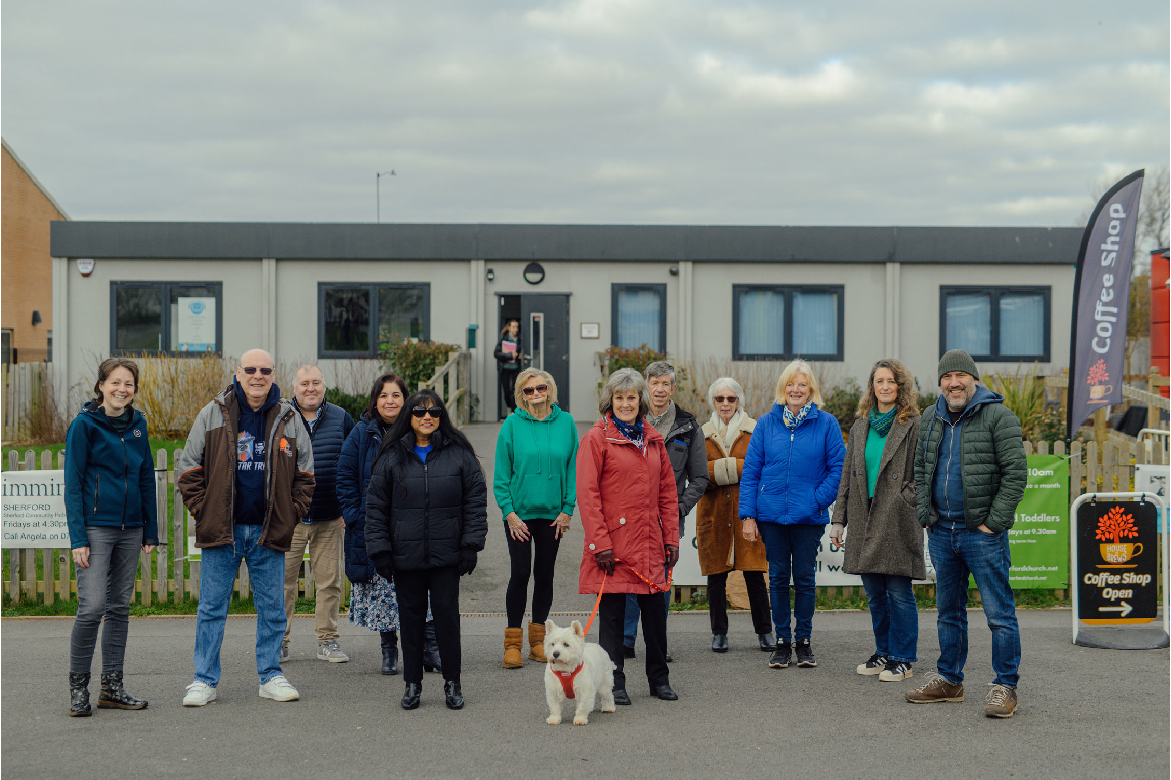A group of eleven people standing outside in front of a single-story building, some wearing jackets and sunglasses, with a white dog on a leash, and a cafe sign on the right side of the image.