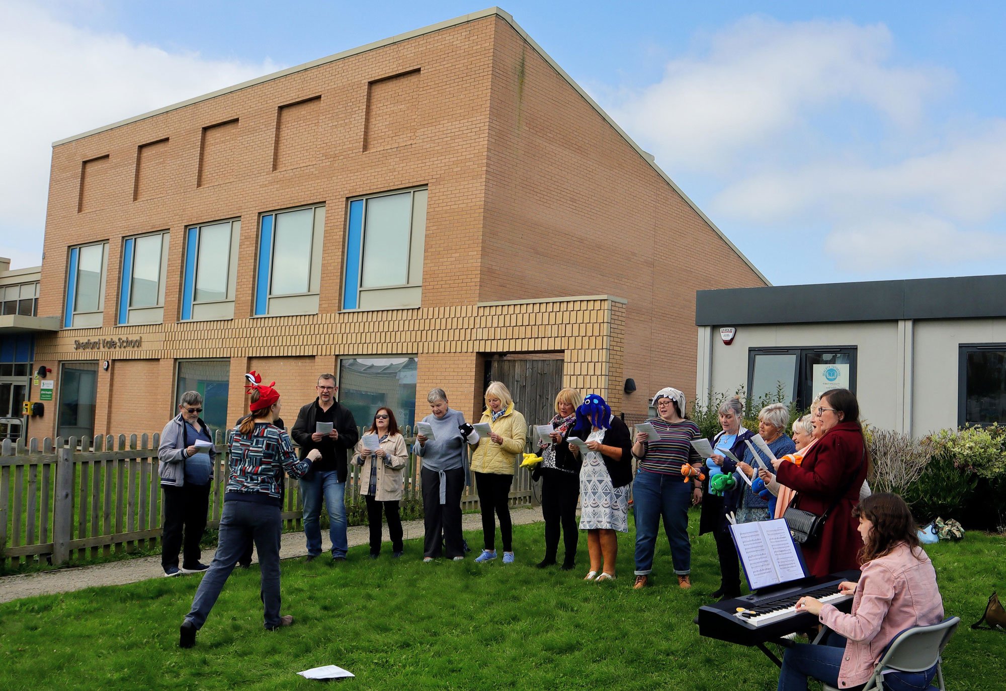 Group of adults singing outdoors in front of a school building, with a woman playing the keyboard.
