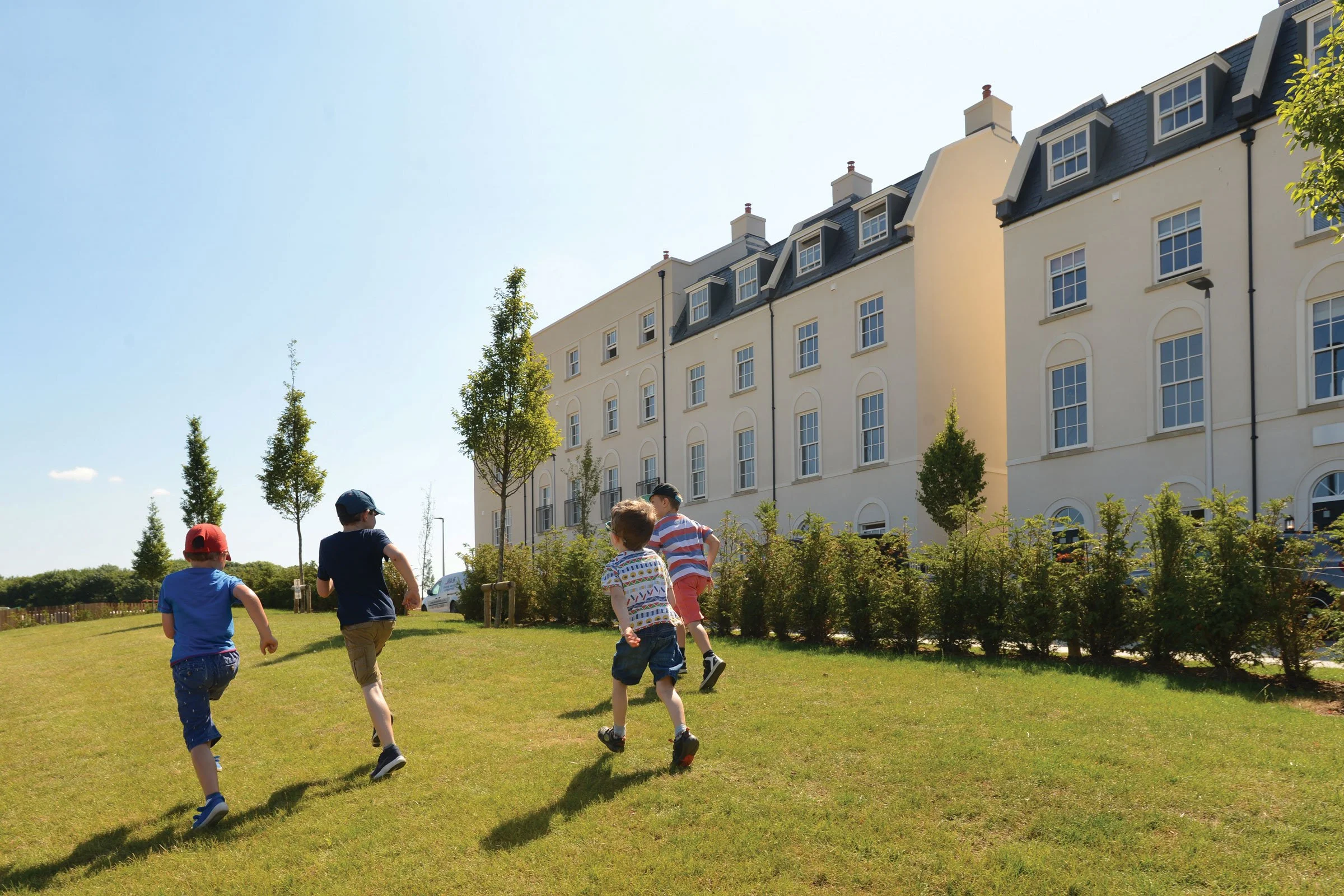 Four children running on a grassy hill near a white multi-story building with multiple windows and a dark roof, under a clear blue sky.