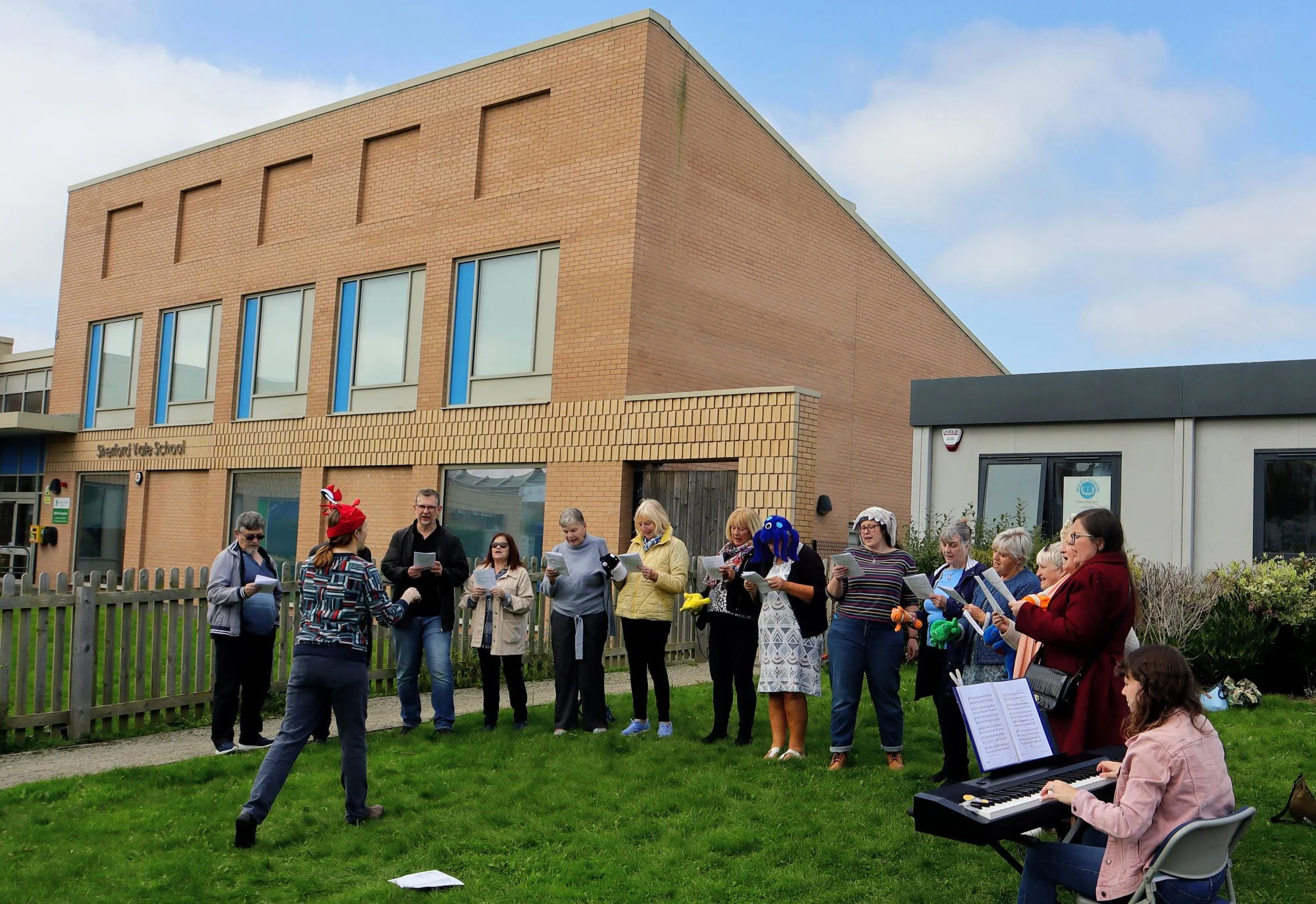 A group of adults singing outdoors in front of a school building, with a person playing piano and a conductor guiding them.