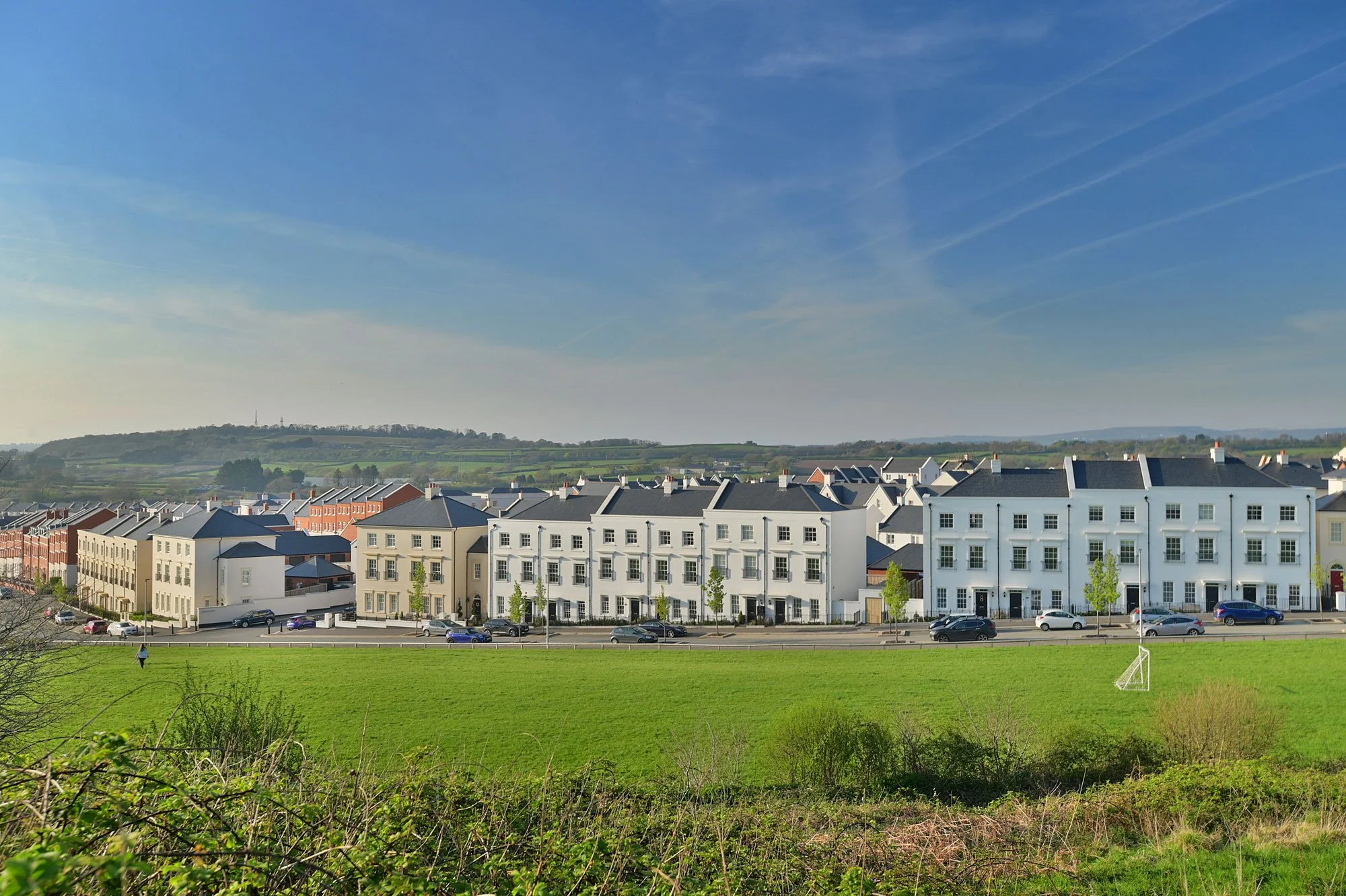 A row of white residential buildings with black roofs along a street, cars parked in front, green grass in the foreground, and hills in the background under a blue sky with contrails.
