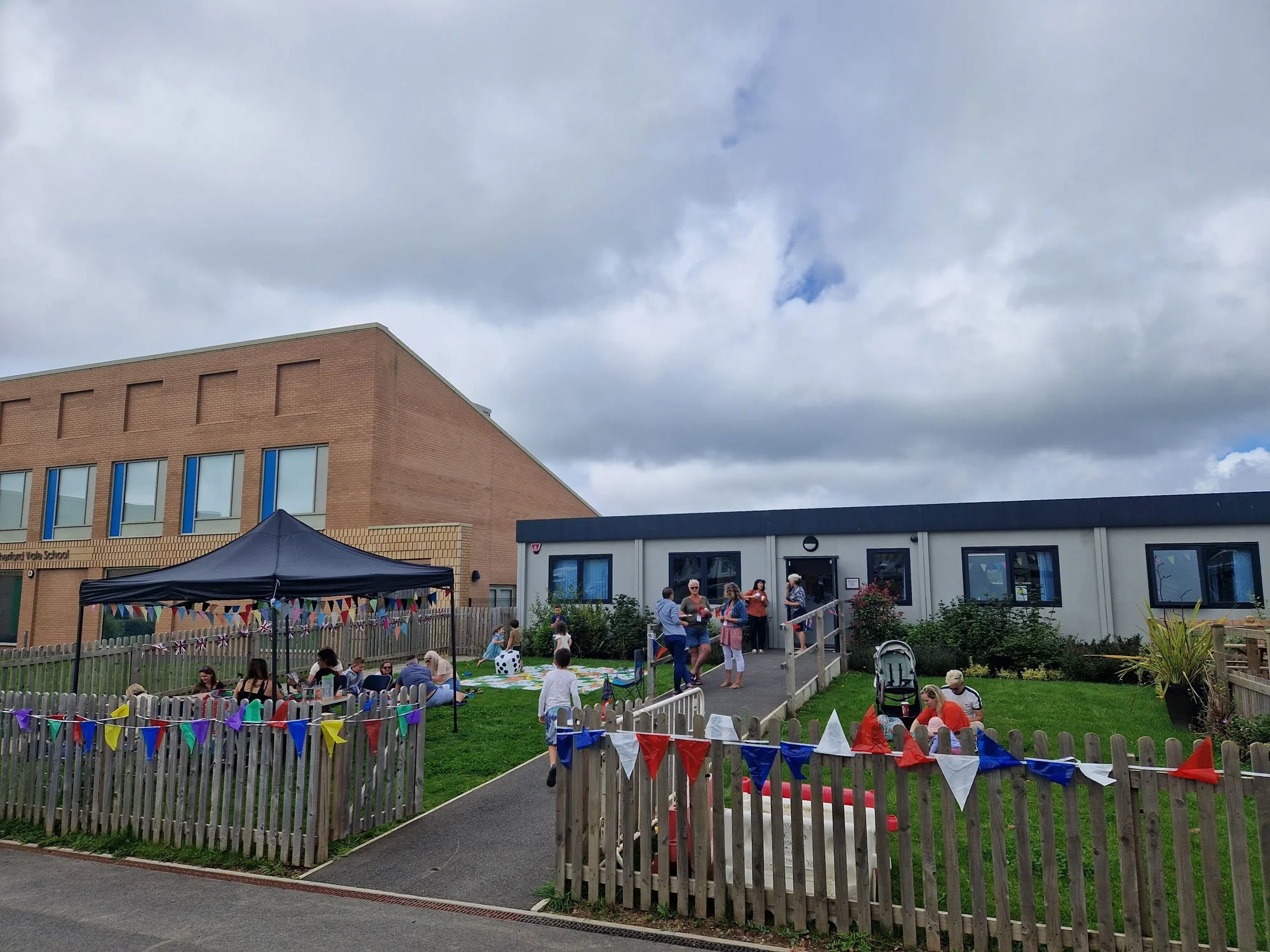 A school event taking place outside a building with a garden, decorated with colorful bunting, with children and adults gathered around, some sitting under a tent, and others walking or standing near the entrance.