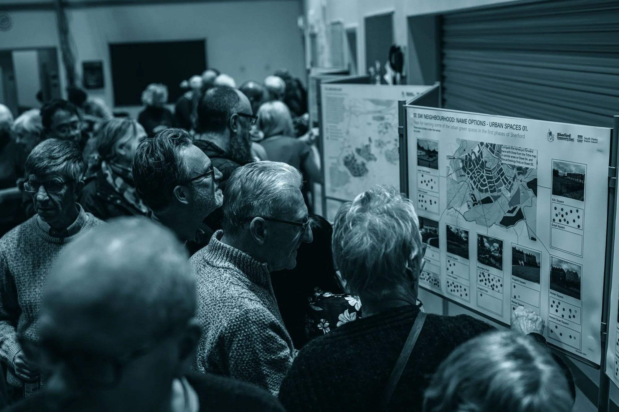 A large group of people attending a presentation or community meeting, looking at display boards with maps and information about urban spaces in Sherford.