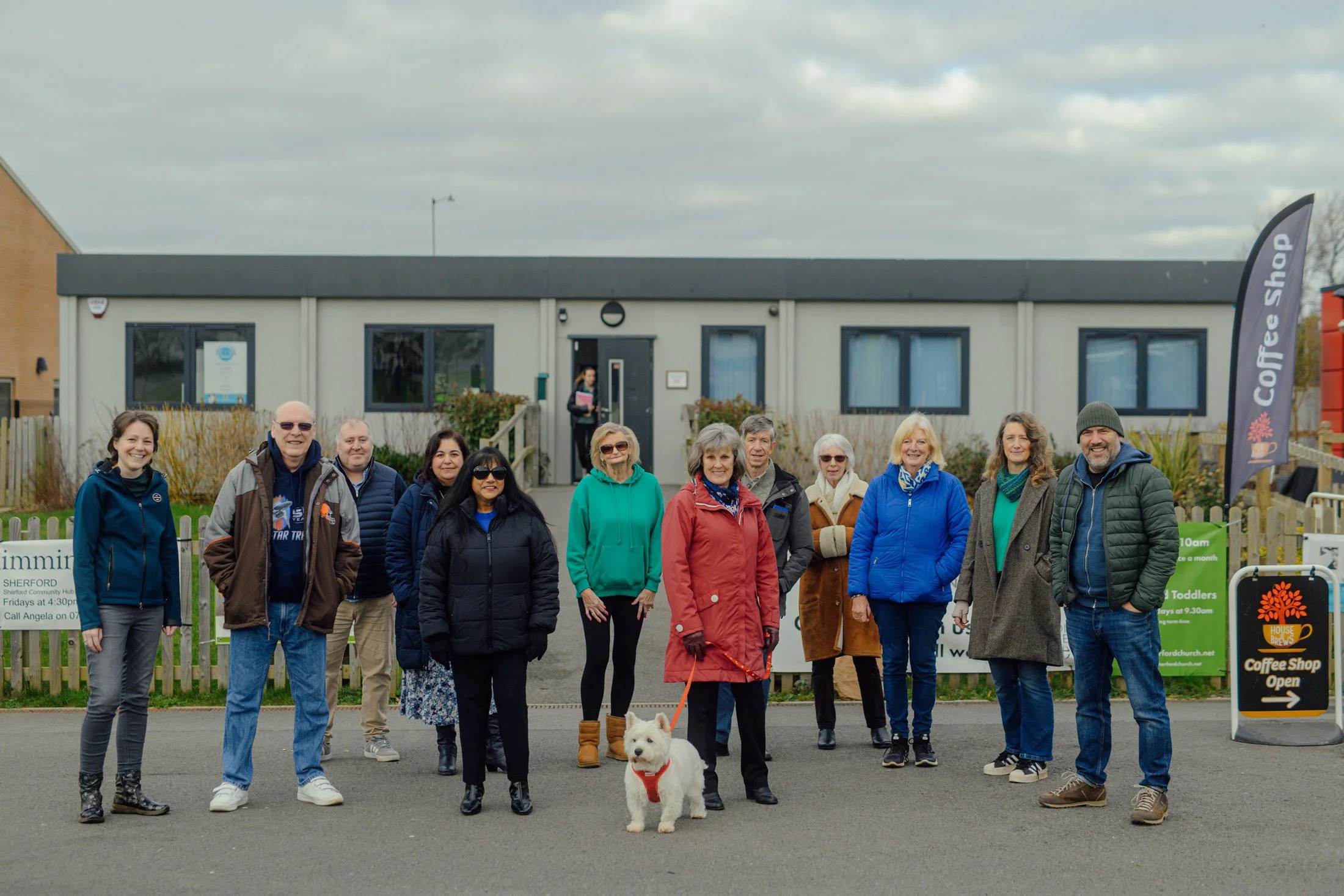 A group of twelve people, including a dog on a leash, standing outside a building with signs for a coffee shop and a swimming pool, under a cloudy sky.