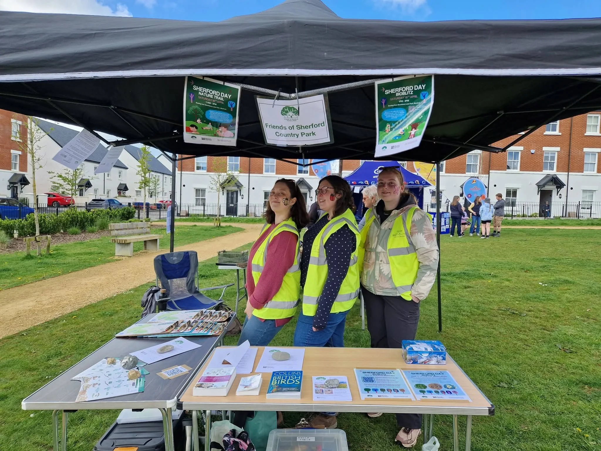 Three adult volunteers, standing under a gazebo, in hi-vis vests at an outdoors community event.  They have a table with leaflets and information in front of them.