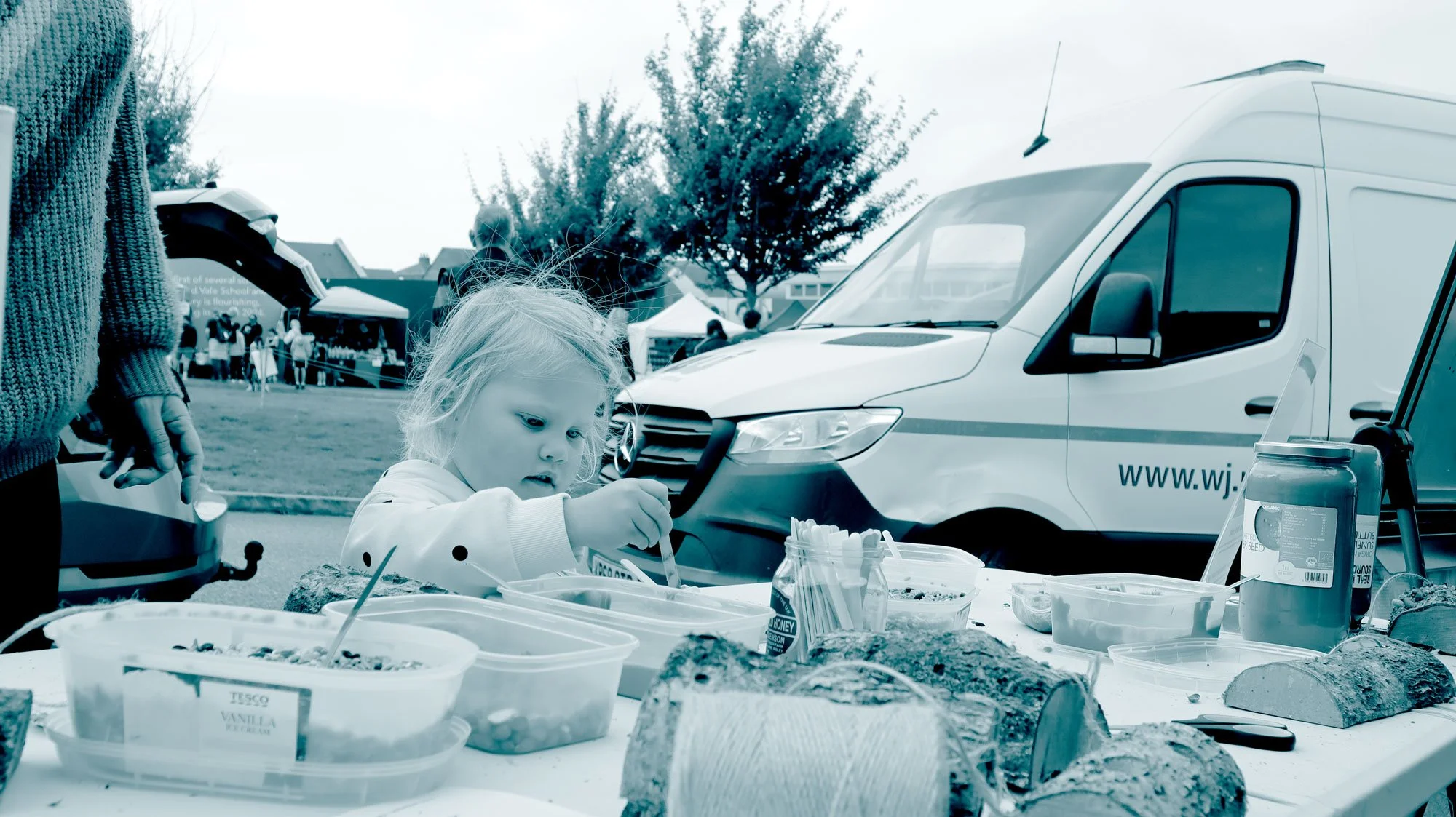 A young girl with blonde hair at a table outdoors during a community event or market, with various food containers, bread, and a jar of jam on the table, and parked vehicles in the background.