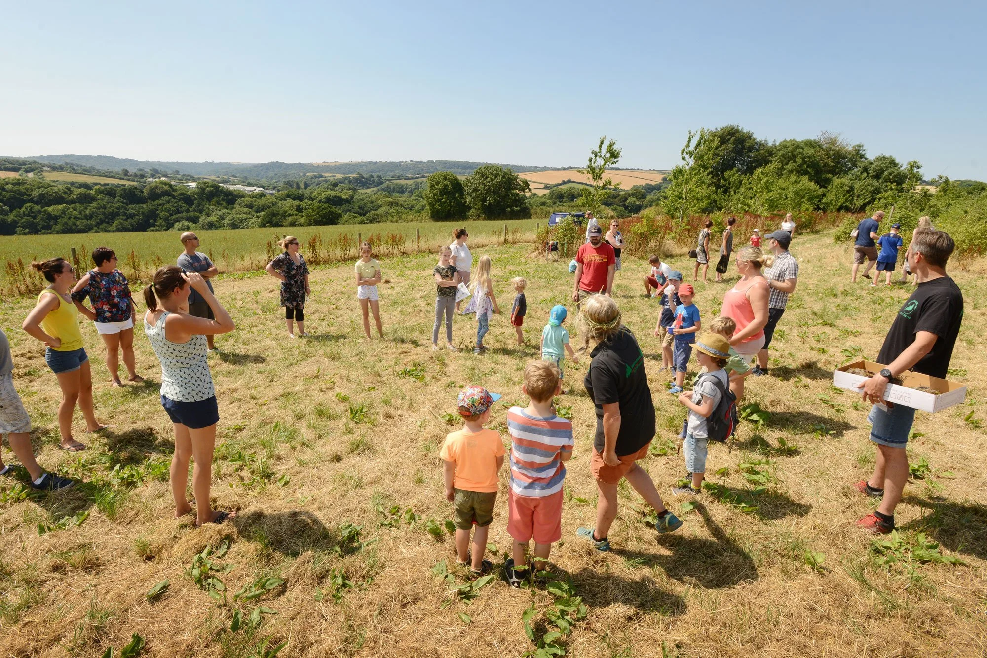 A group of people, including adults and children, gathered outdoors on a sunny day in a grassy field surrounded by trees and rolling hills, appearing to participate in an organized event.