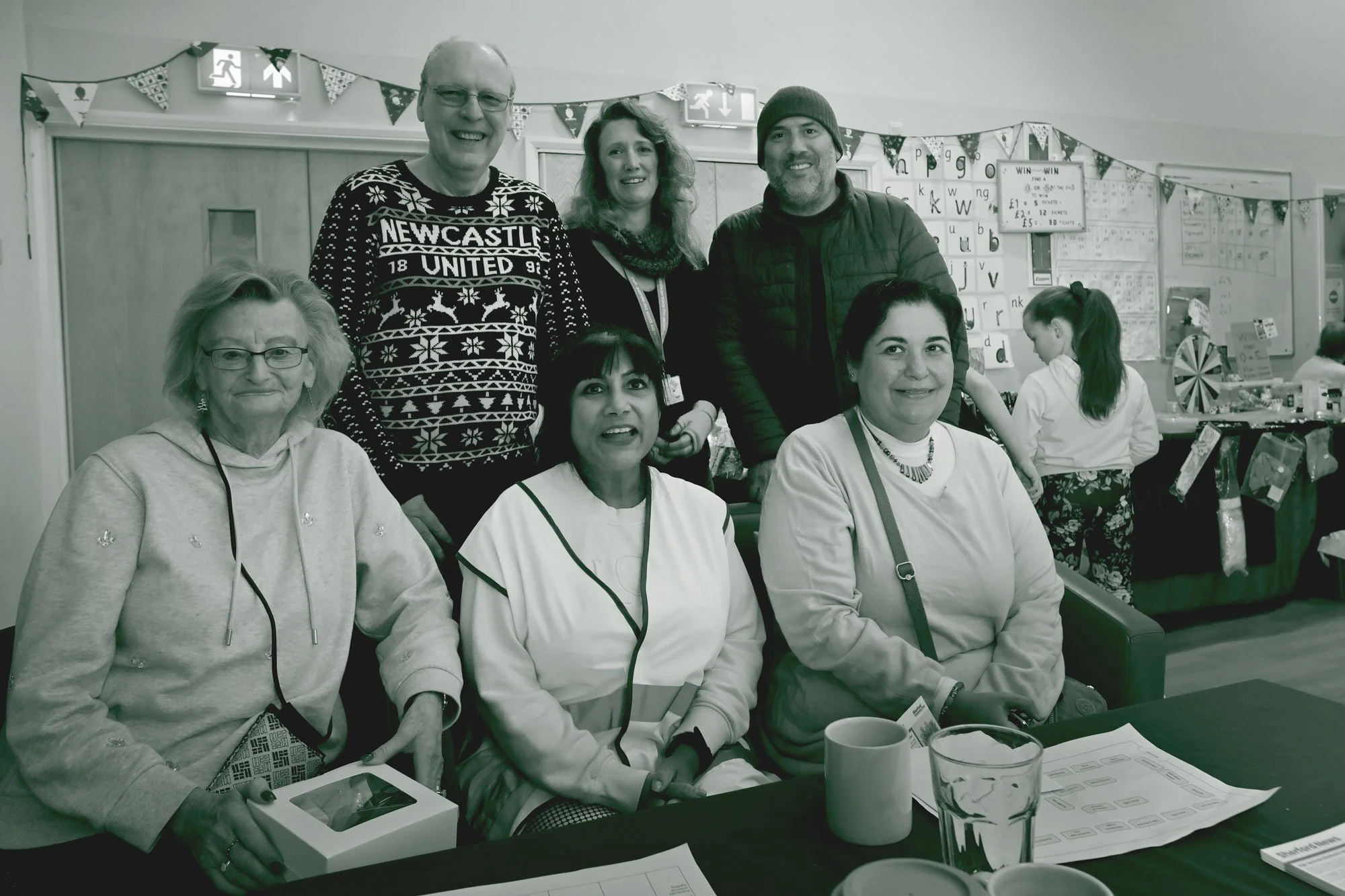 Group of six people, three sitting and three standing, in a festive indoor setting with Christmas decorations and a Christmas sweater, smiling at the camera.