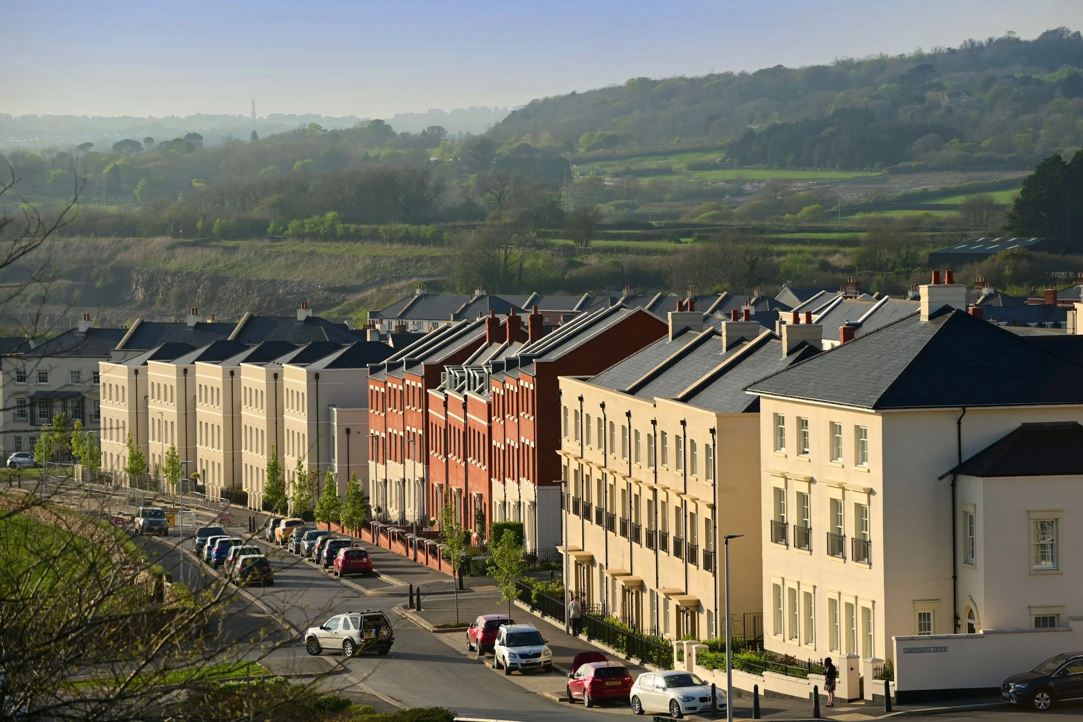 Row of modern residential buildings with parking lot and cars in the foreground, green hills and trees in the background.