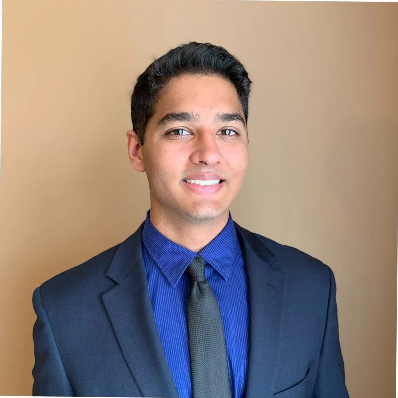 A young man in a dark suit, blue shirt, and gray tie, smiling and standing against a beige background.