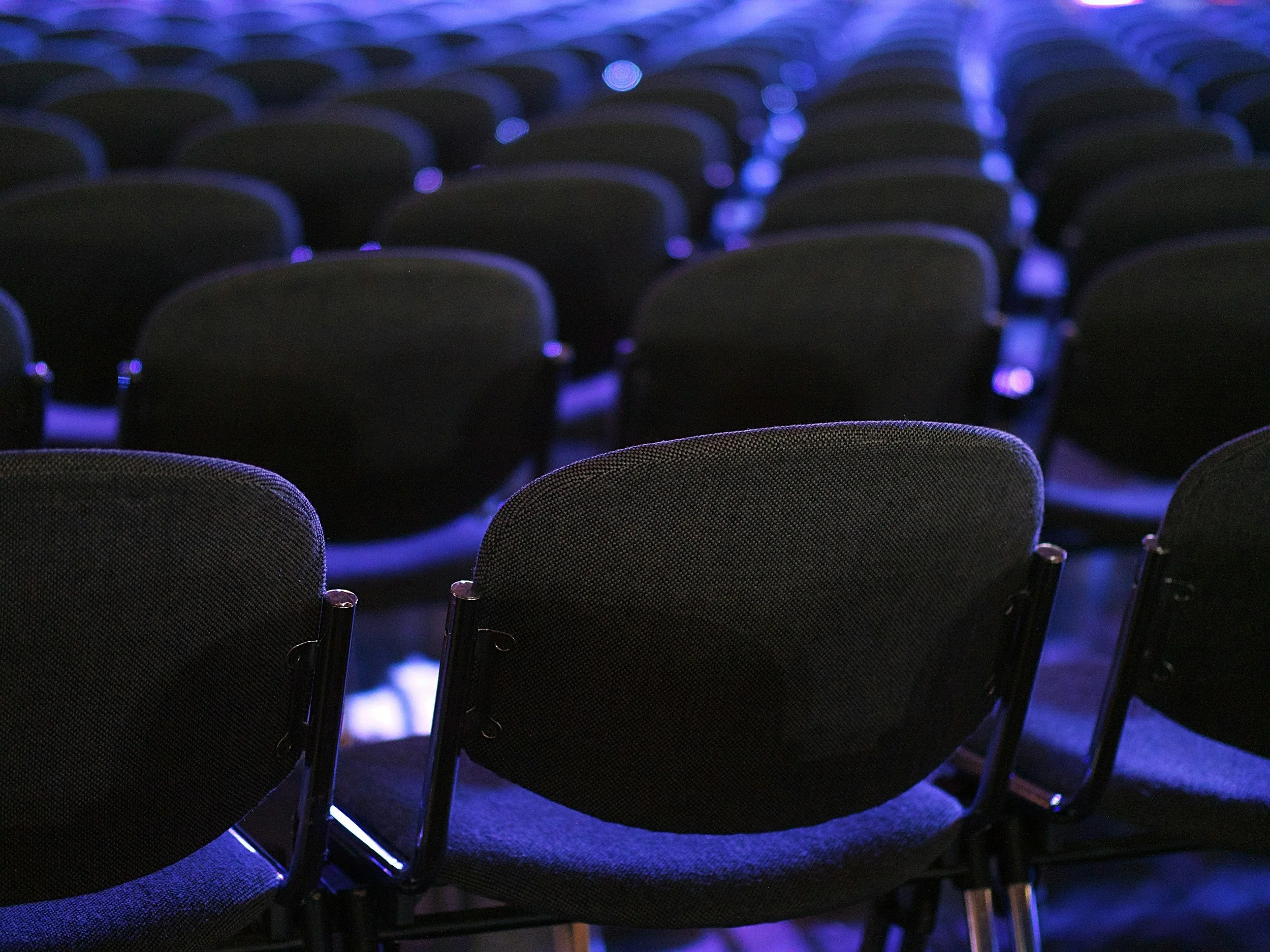 Rows of empty black chairs illuminated by blue and purple lighting, arranged in a theater or conference hall setting.