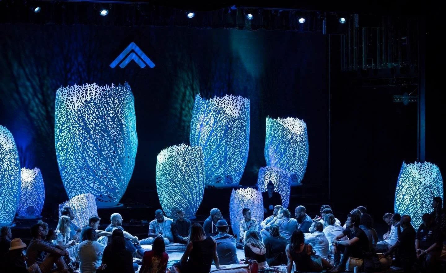 People seated on the floor in front of large illuminated art installations resembling glowing coral or shells in a dark room, with a logo at the top center.