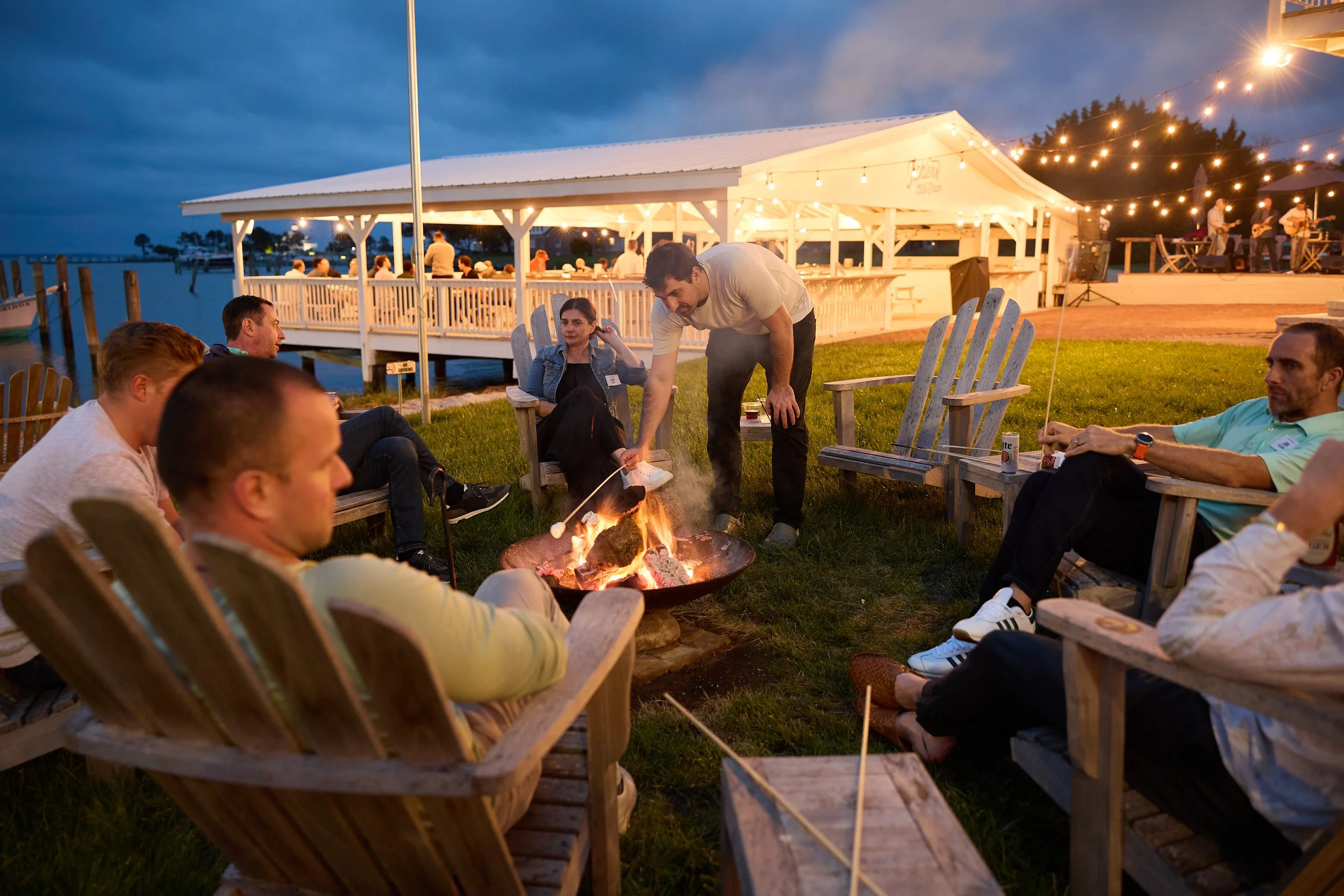 Group of people sitting outdoors around a fire pit at dusk, enjoying a barbecue with a waterfront view and string lights overhead, while a man roasts marshmallows.