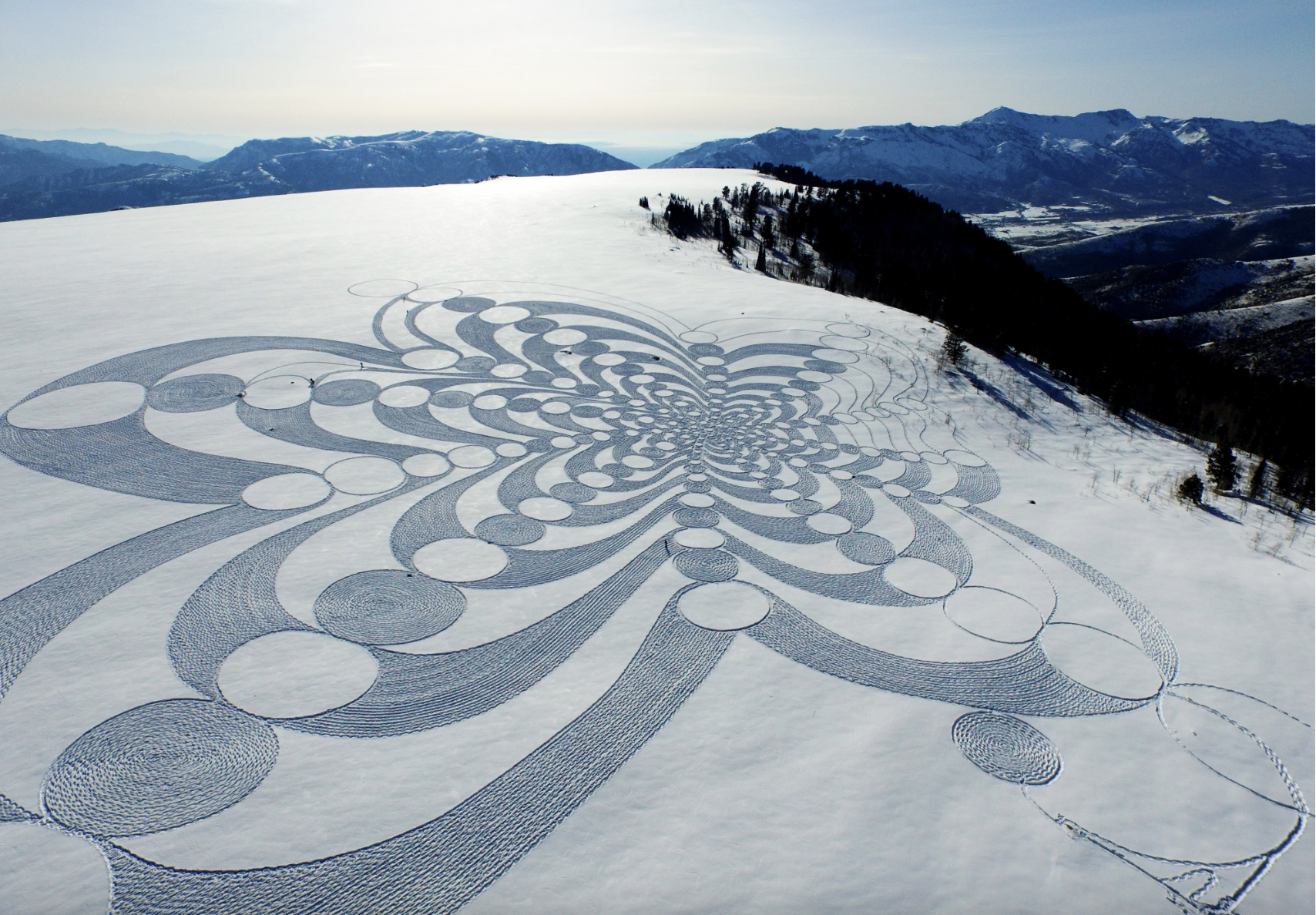 Snow-covered landscape with a large, intricate geometric snow art pattern featuring circles and flowing lines on a hillside, with mountains and trees in the background.