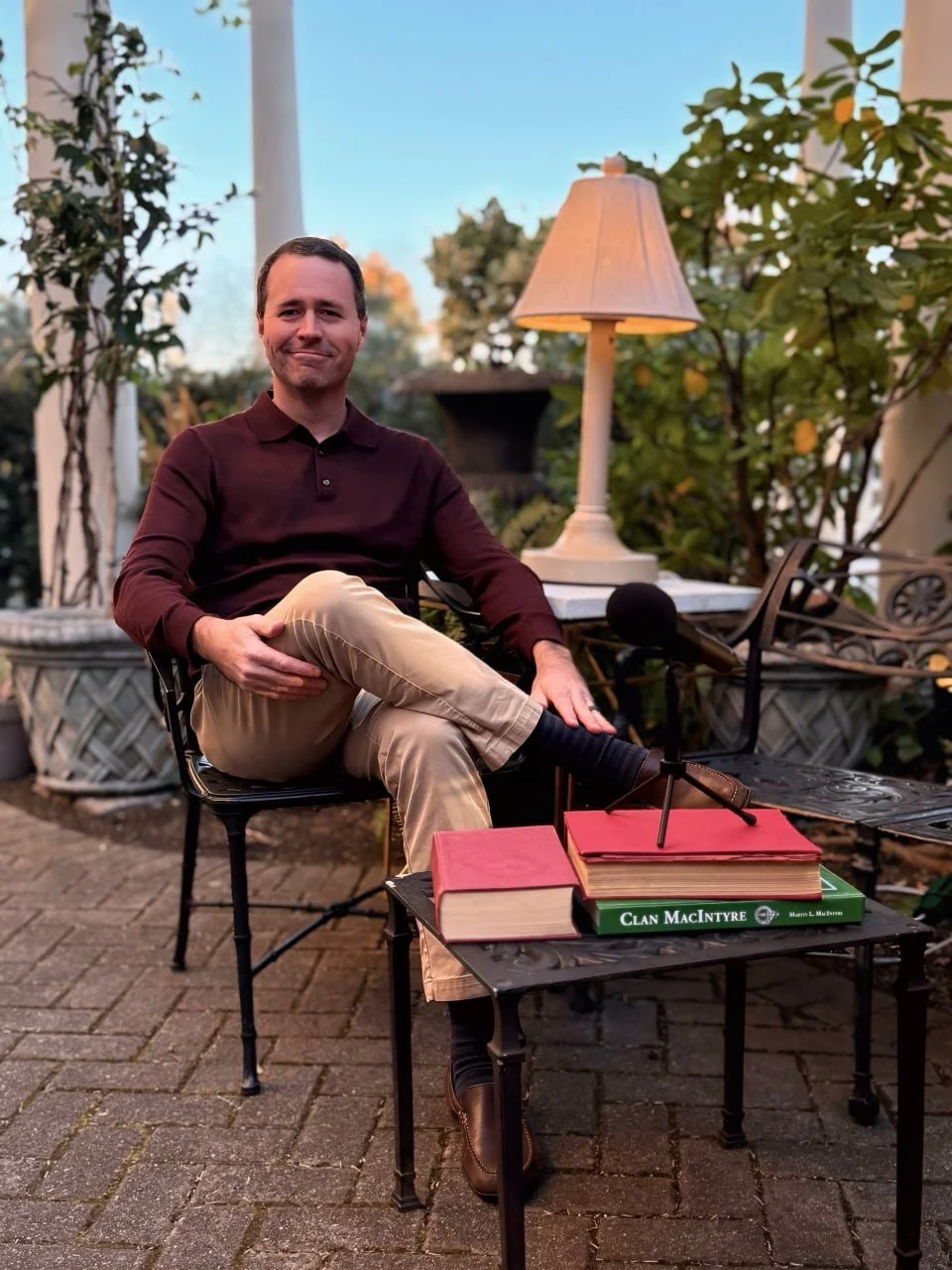 A man sitting outdoors at a table with a microphone, books including one titled "Clan MacIntyre", a lamp, and a decorative bench, with trees and a blue sky in the background.