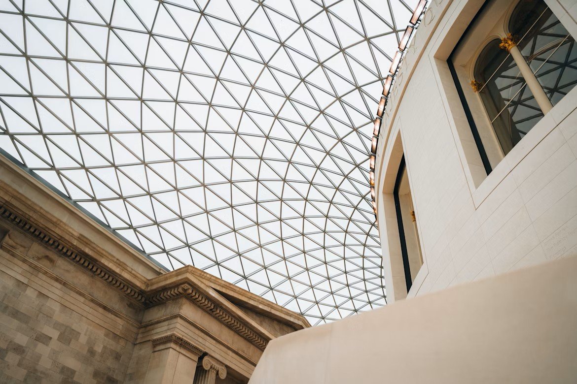 Interior view of a building showcasing a glass and metal lattice ceiling, with classical architecture elements like stone walls and ornate details, alongside modern design features.