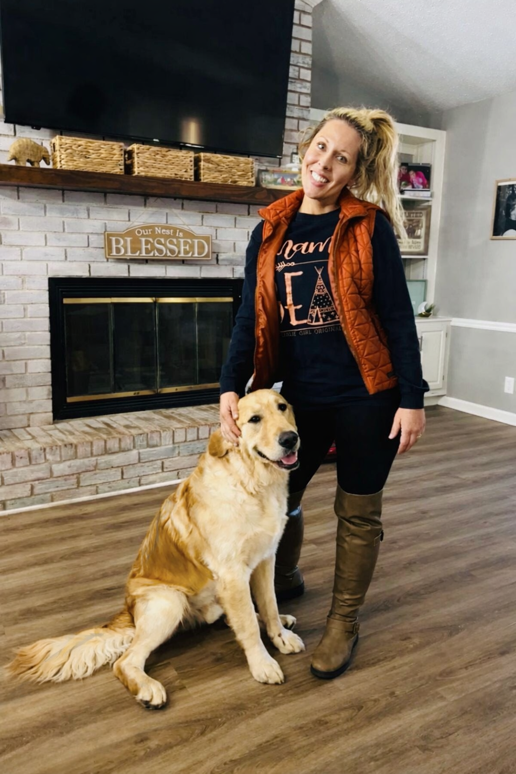 A woman with blonde hair tied back, wearing a black t-shirt with 'Mama' printed on it, a brown quilted vest, and brown riding boots, is standing indoors next to a golden retriever dog. She is smiling and holding her hand on the dog's head. The background features a brick fireplace with a sign that says 'Our Nest Is Blessed', a flat-screen TV mounted on the wall, and a white shelving unit with books and pictures.