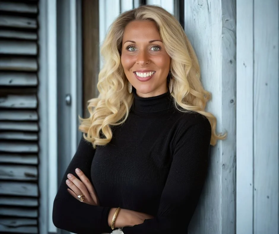 A woman with long blonde curly hair, wearing a black turtleneck, smiling and standing with arms crossed against a wooden wall.