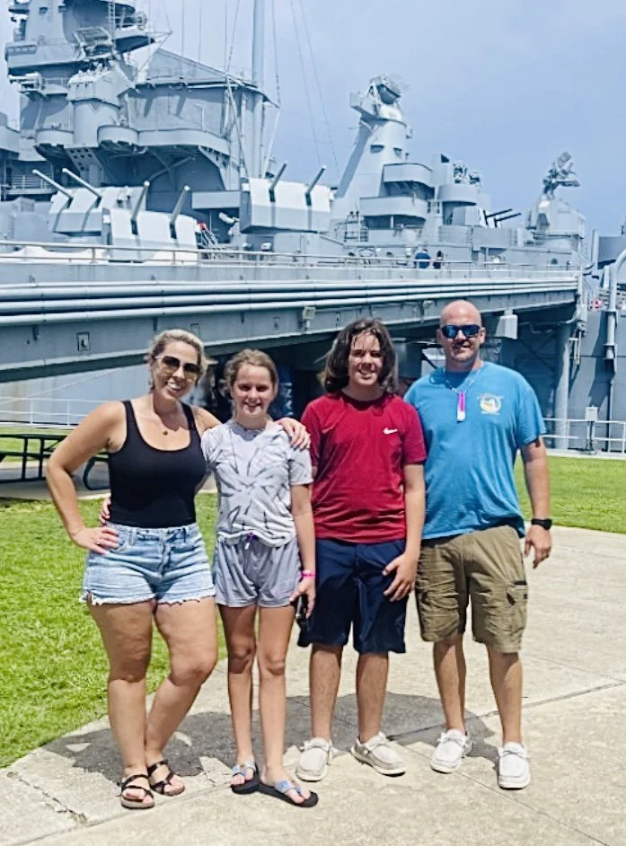 Family of four standing in front of a large battleship on a sunny day.