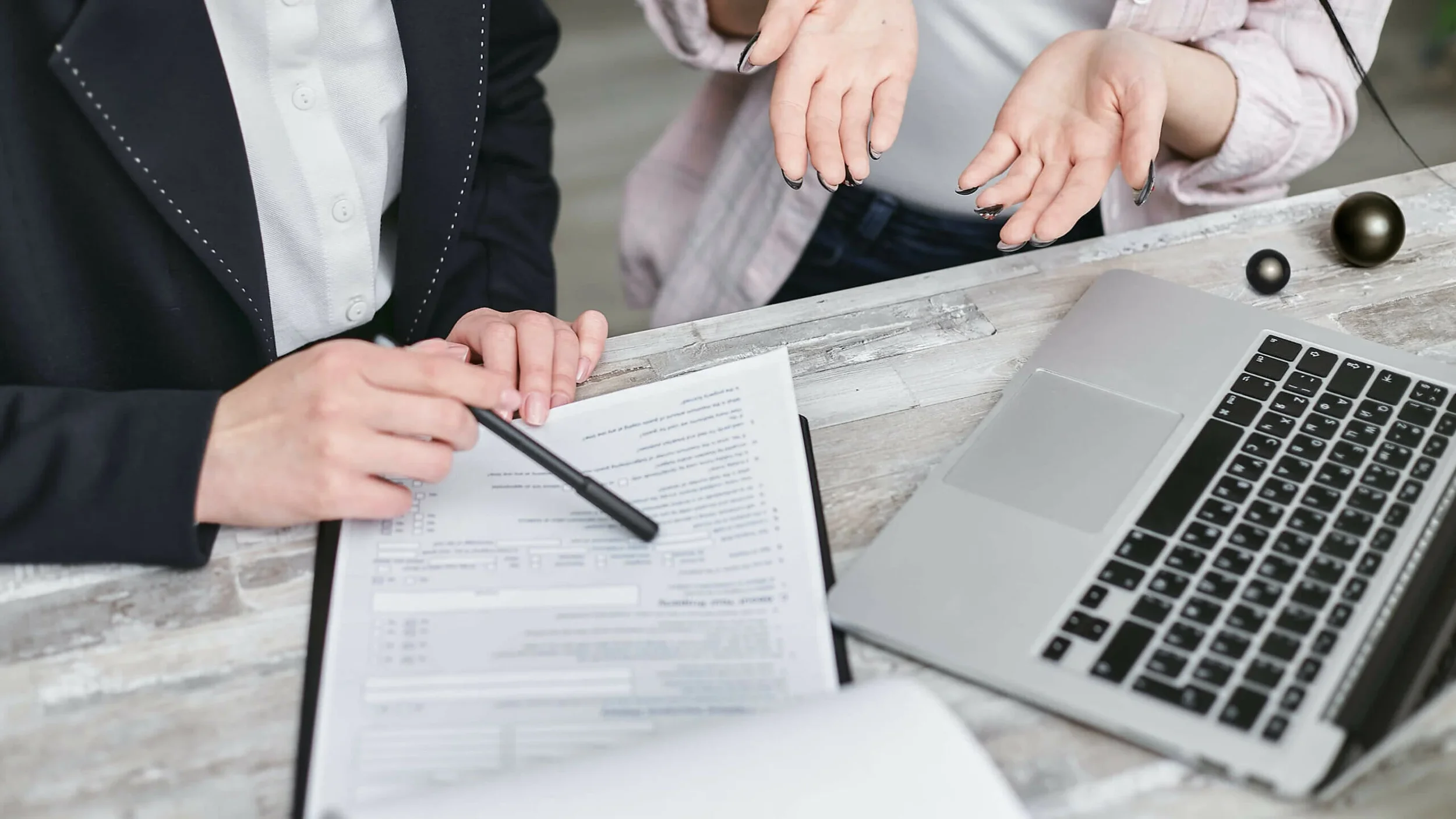 Two people discuss financial documents at a wooden table with a laptop and black decorative balls.