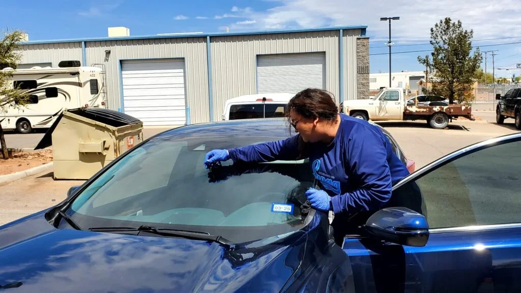 A person inspecting or cleaning the windshield of a blue car outdoors in a parking lot, with industrial buildings and other vehicles in the background.