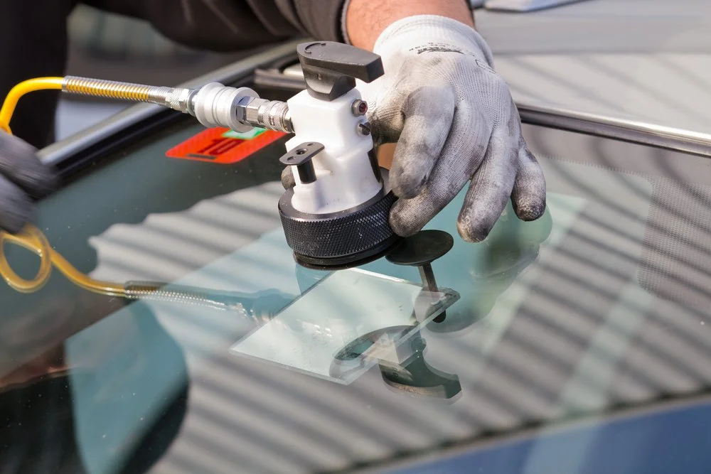 A person wearing a glove uses a precision tool on a vehicle's windshield, likely for glass installation or repair.