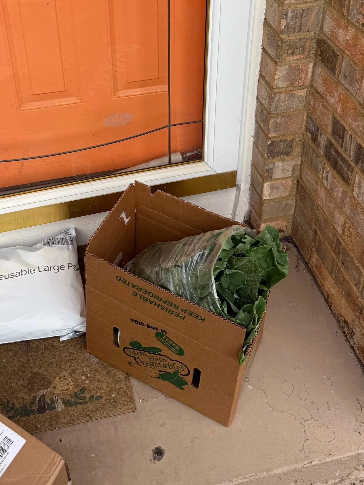 A cardboard box filled with leafy green vegetables placed on a porch or doorstep near a glass door with a brick wall on the side.