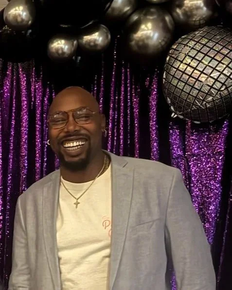 A smiling man with glasses and a gold chain, dressed in a light gray blazer and white shirt, standing in front of a purple sequin backdrop with large silver balloons and a disco ball.