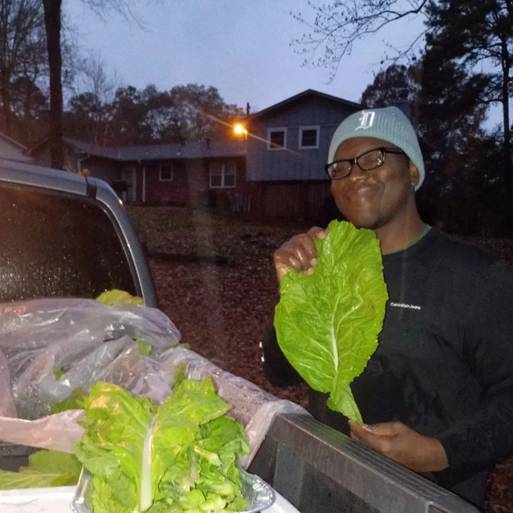 A man holding a large florida broadleaf mustard green leaf , smiling and standing outdoors during dusk, next to a car with additional greens in the trunk.