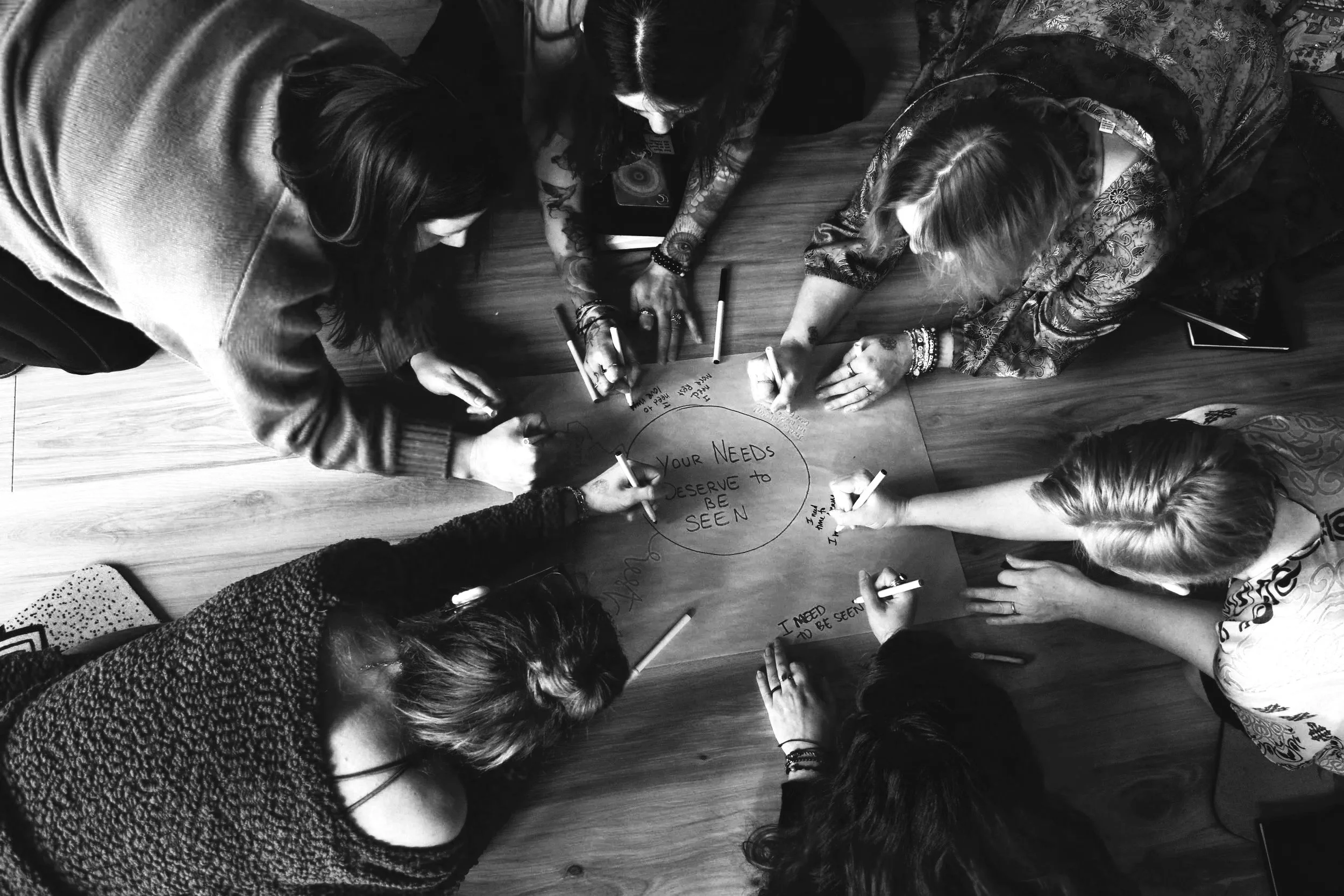 Top-down view of six women gathered around a table, writing on a large sheet of paper with the message 'Your Needs Deserve to be Seen' circled at the center.