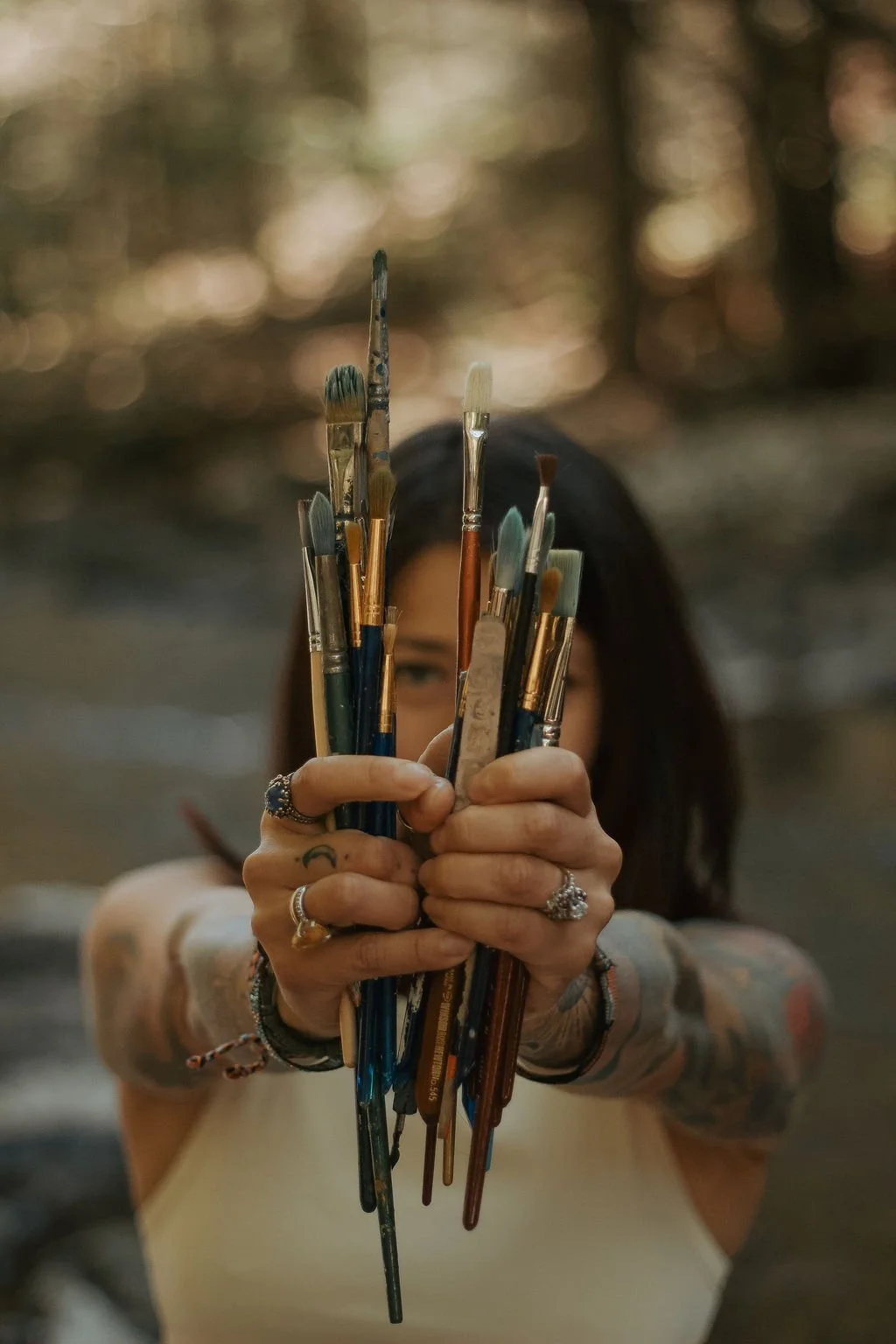A person holding a bunch of paintbrushes, with their face partially obscured behind them, outdoors with a blurred background of trees.