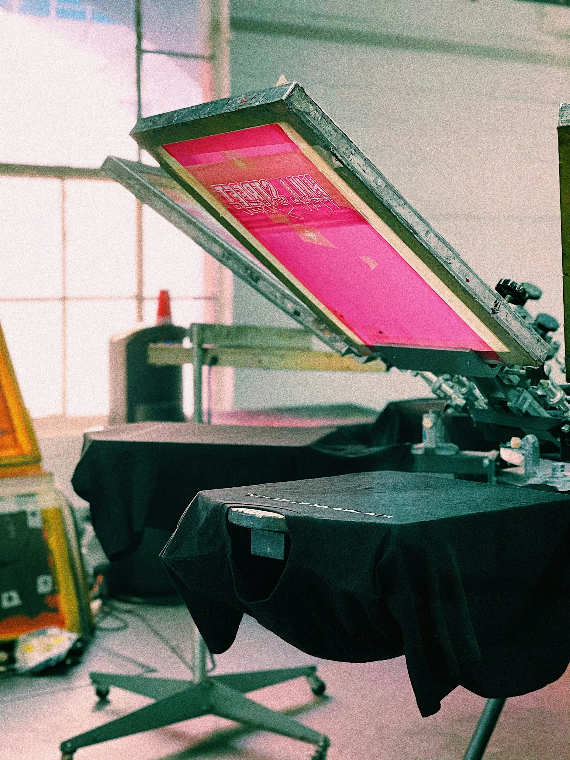 Screen printing press with a pink screen and black fabric on the table, in a bright workspace with large windows.