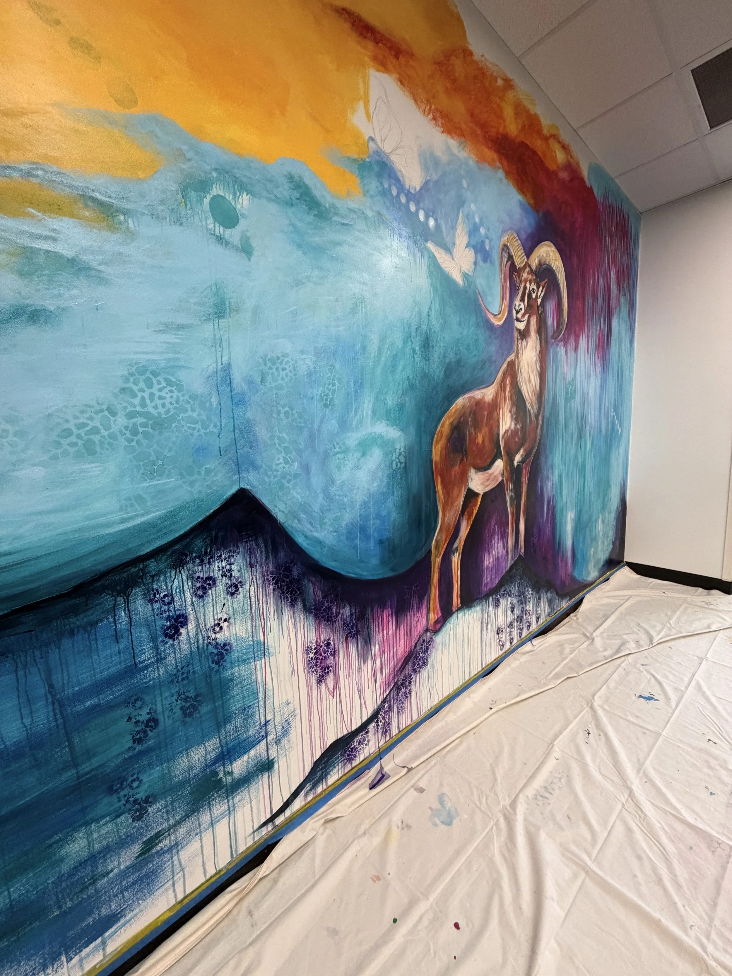 Colorful mural featuring a goat with large horns, standing on a mountain with blue water and sky background. The mural is being painted on a wall, with white and purple paint splatters on the floor below.