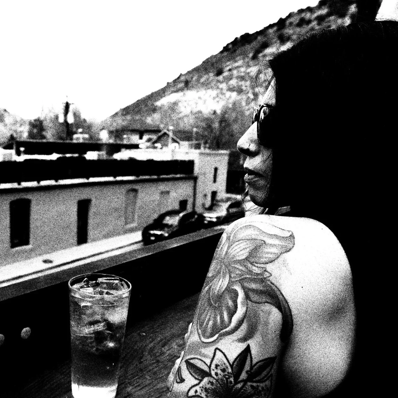 A black and white photo of a woman with tattoos on her shoulder and arm, wearing glasses, sitting at a table with a drink that contains ice. She is looking to the right, and there is a scenic view of mountains and buildings in the background.