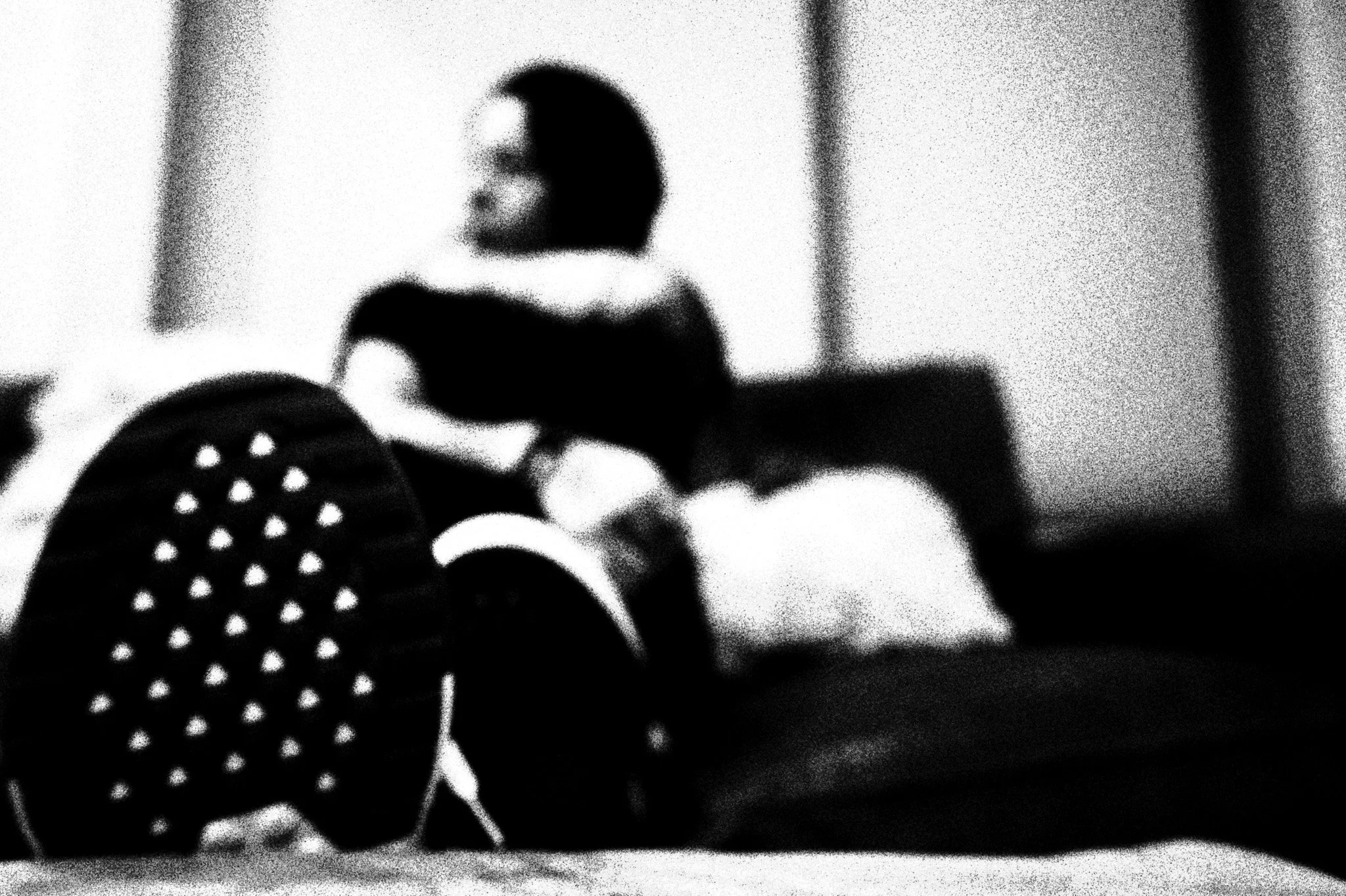 A blurry, black-and-white photo of a person sitting in a room, with a perforated chair visible in the foreground.