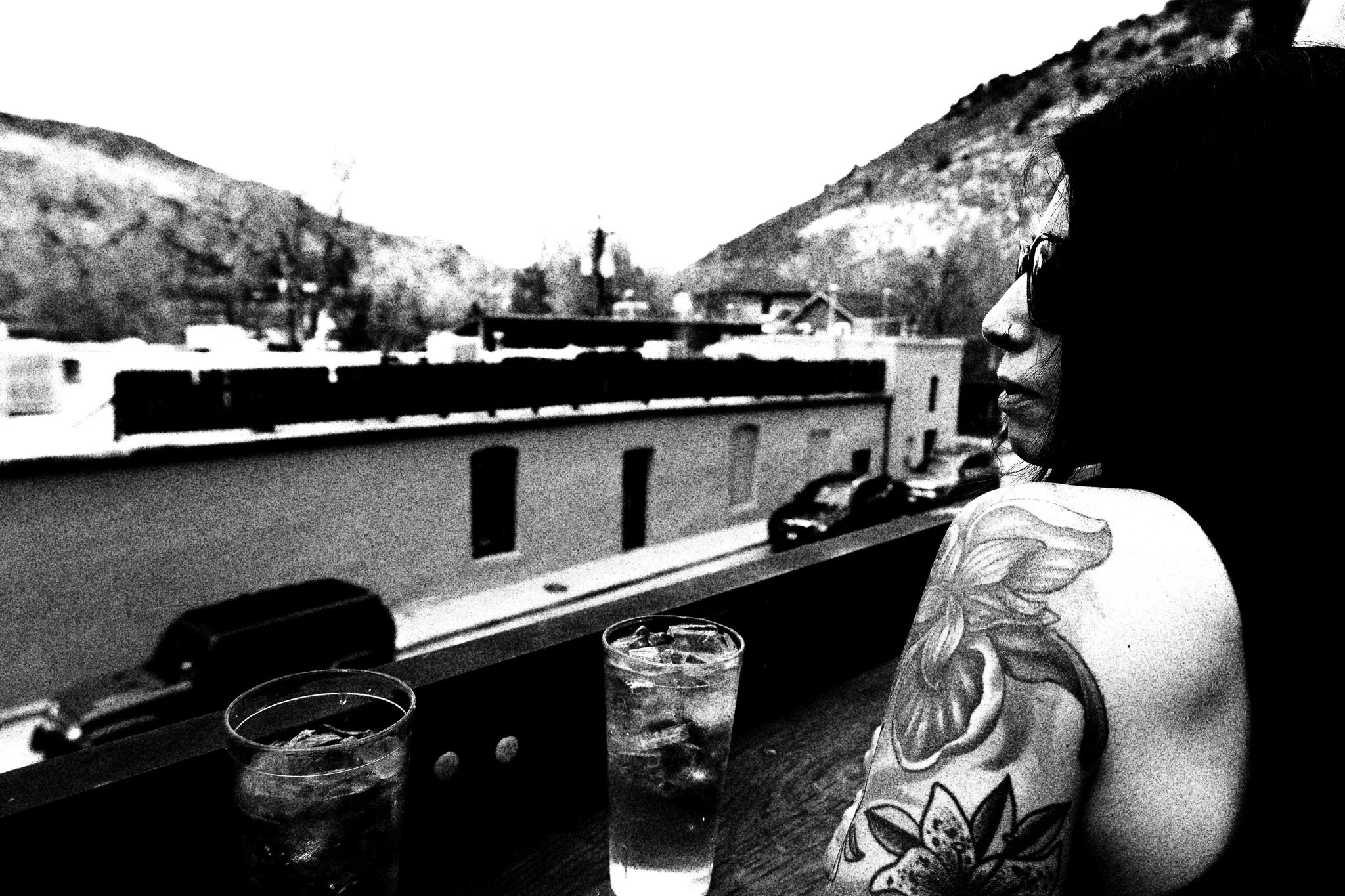 A woman with long dark hair and sunglasses sitting on a balcony, looking towards the street. She has a large tattoo of a lion's face and a flower on her shoulder. There are two glasses of iced drinks on the railing.