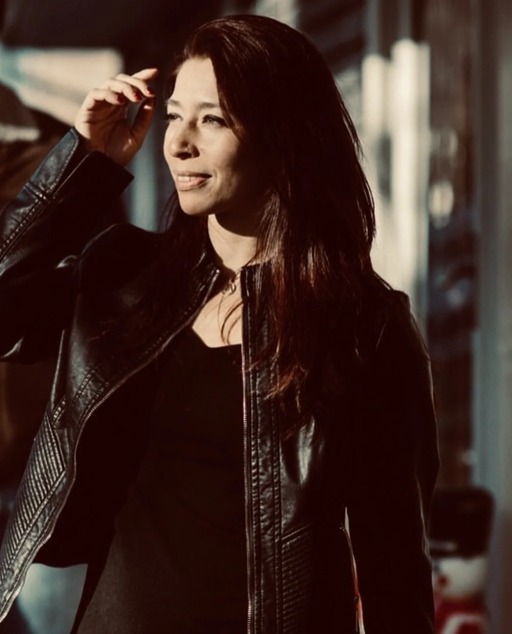 A woman with long dark hair wearing a black leather jacket, standing in a warmly lit room.