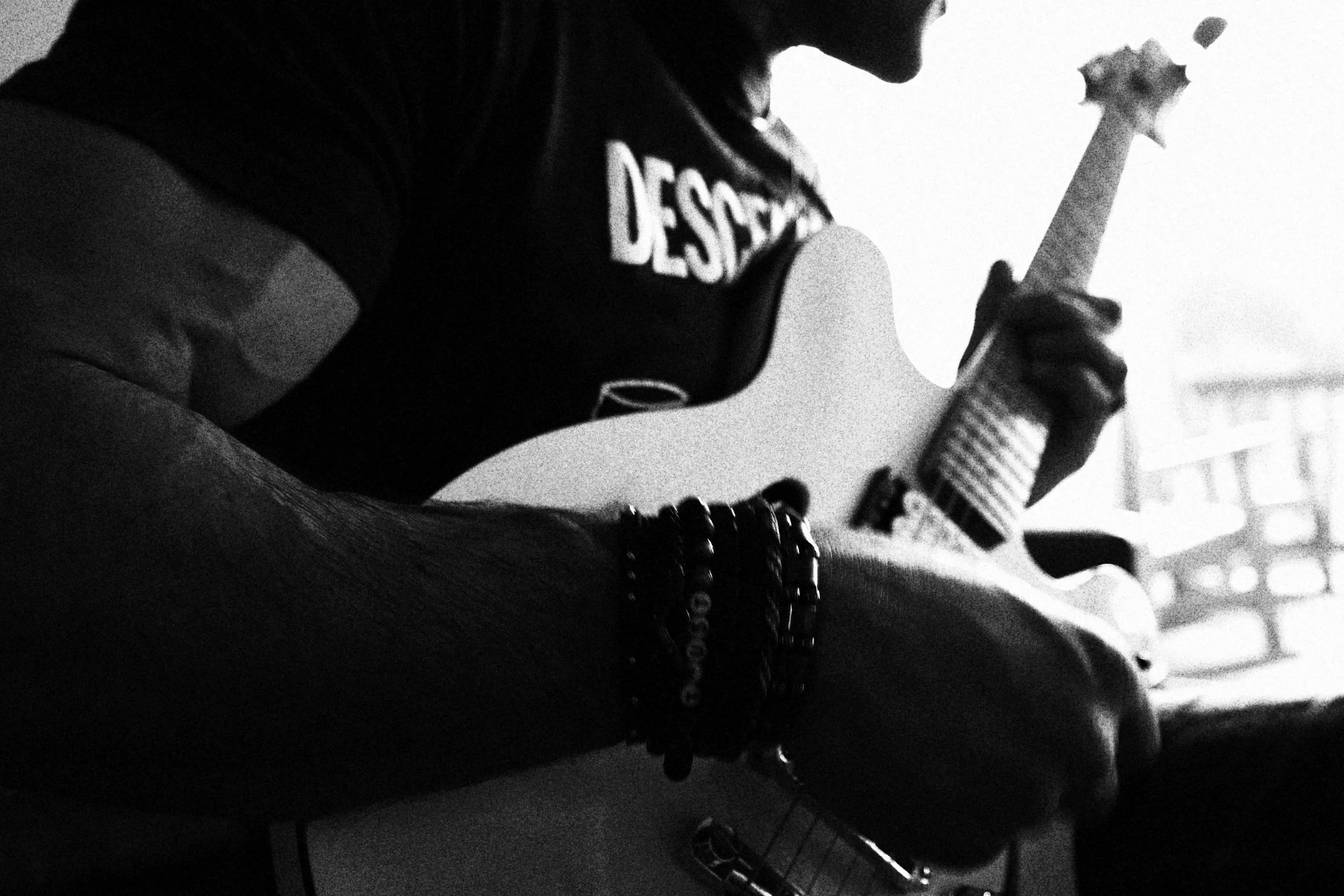 Close-up black-and-white photo of a person playing an electric guitar with bracelets on their wrist, wearing a t-shirt with the word 'DESIGN' printed on it.