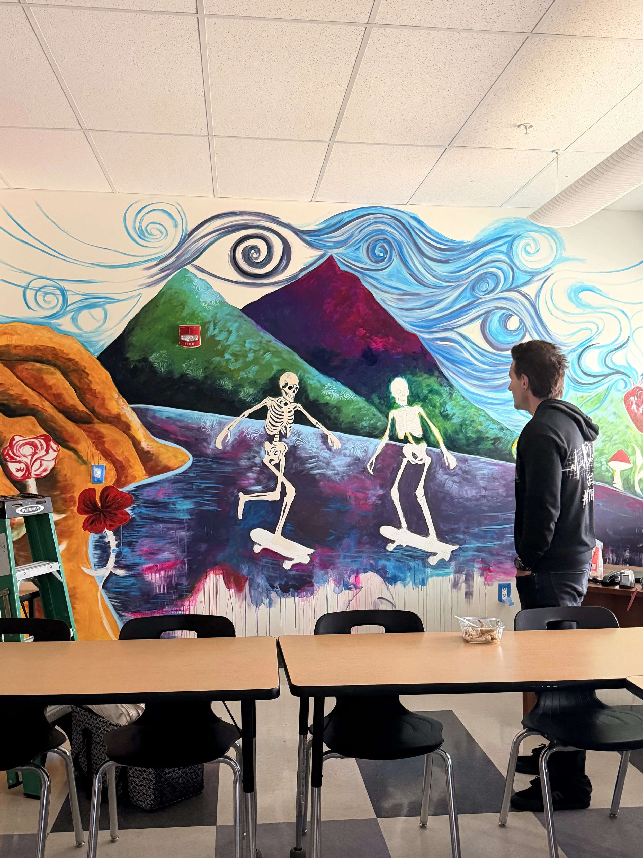 Colorful mural depicting mountains, swirling wind patterns, and skeletal figures skateboarding, with tables and chairs in the foreground.