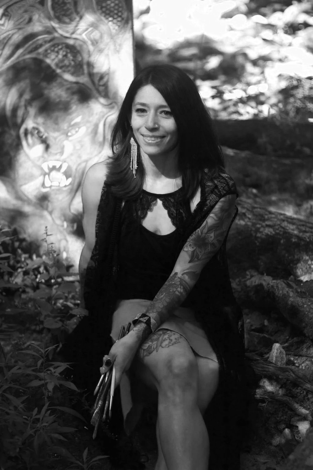 A woman with dark hair, tattoos on her right arm, wearing earrings and a black dress, sitting outdoors near a painted lion mural in a natural setting with trees and rocks in the background.