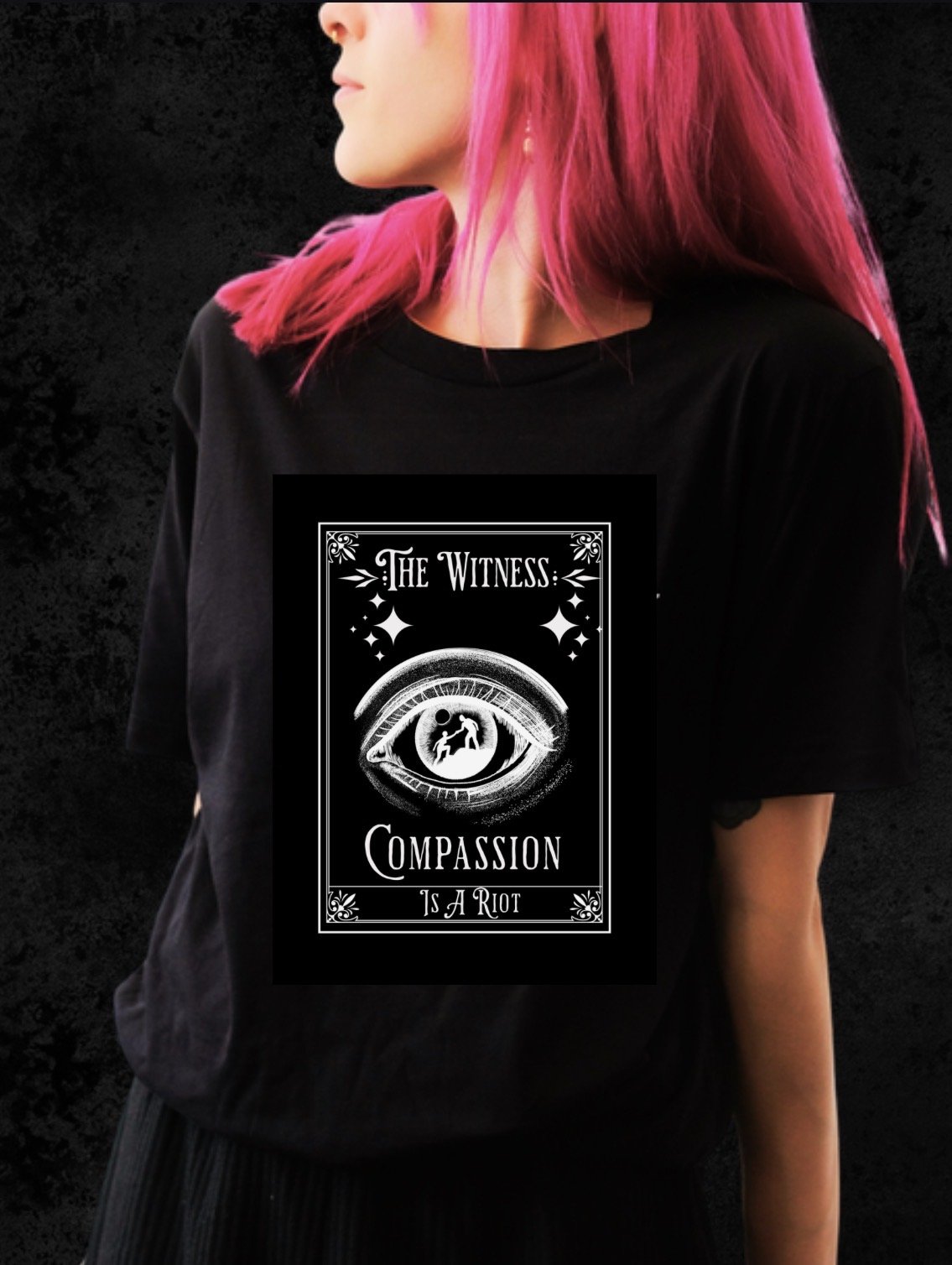 A person with bright pink hair wearing a black T-shirt with a graphic design featuring an eye and the words "The Witness: Compassion is a Riot."