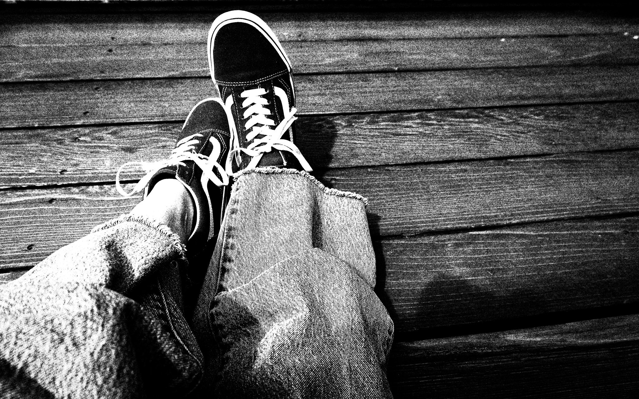 Black and white photo of a person's legs wearing ripped jeans and sneakers, sitting on a wooden surface.