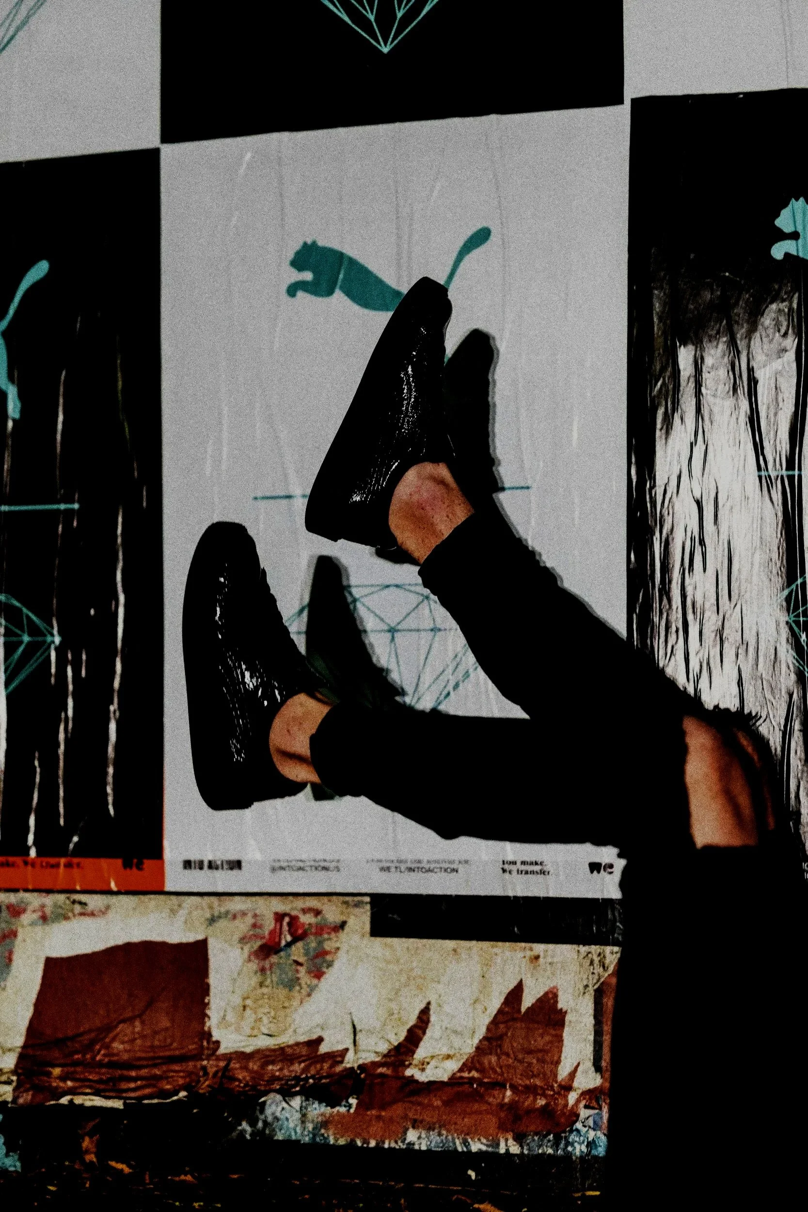 A close-up of a wall covered with various posters and images, including a person standing on a poster with black shiny shoes and black pants, partially visible in the photo. The posters include a panda illustration, some abstract artwork, and a geometric design at the top.