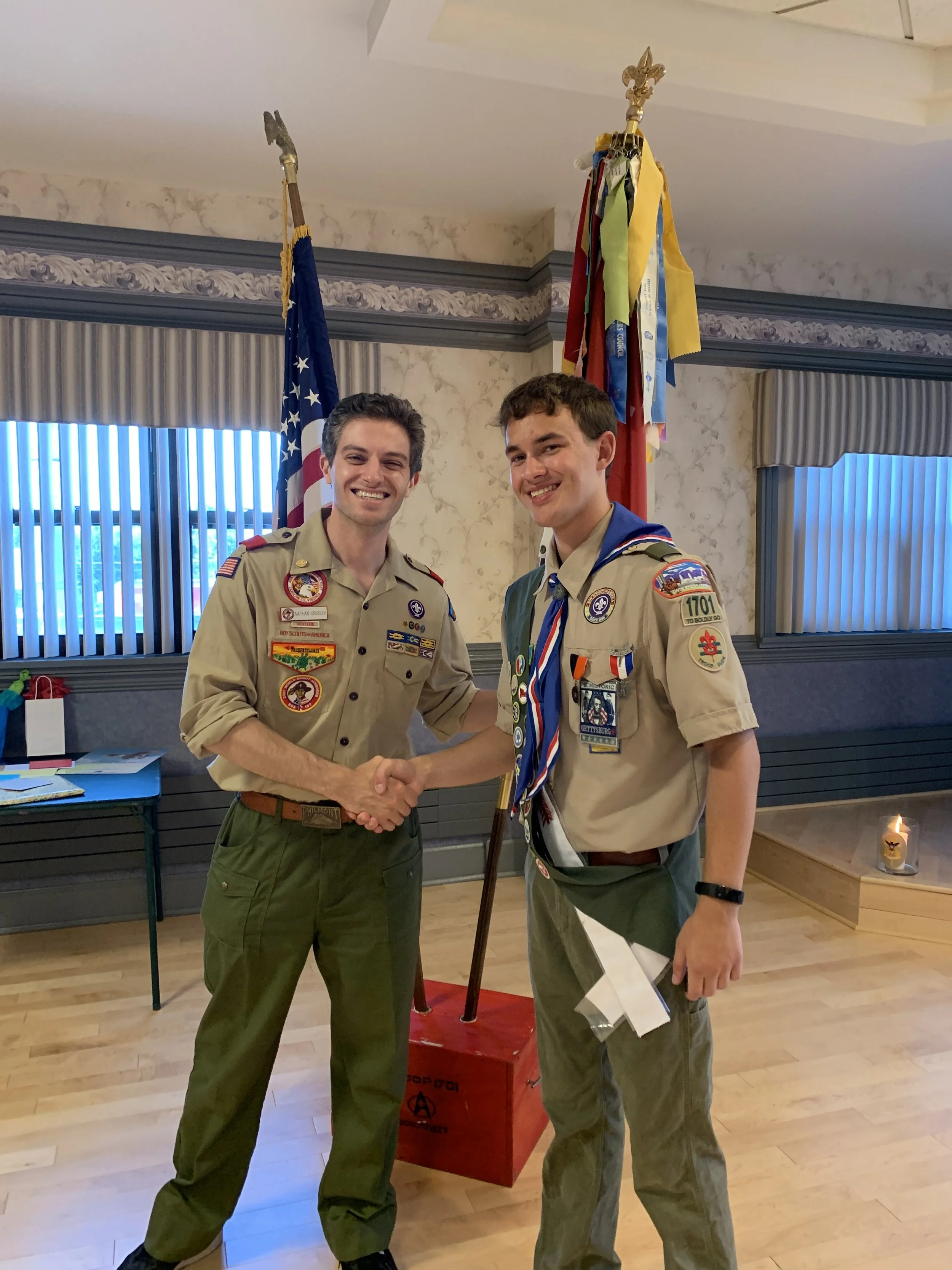Two young men shaking hands, both dressed in Boy Scout uniforms, with awards and patches on their shirts, in a room with flags and scout banners in the background.