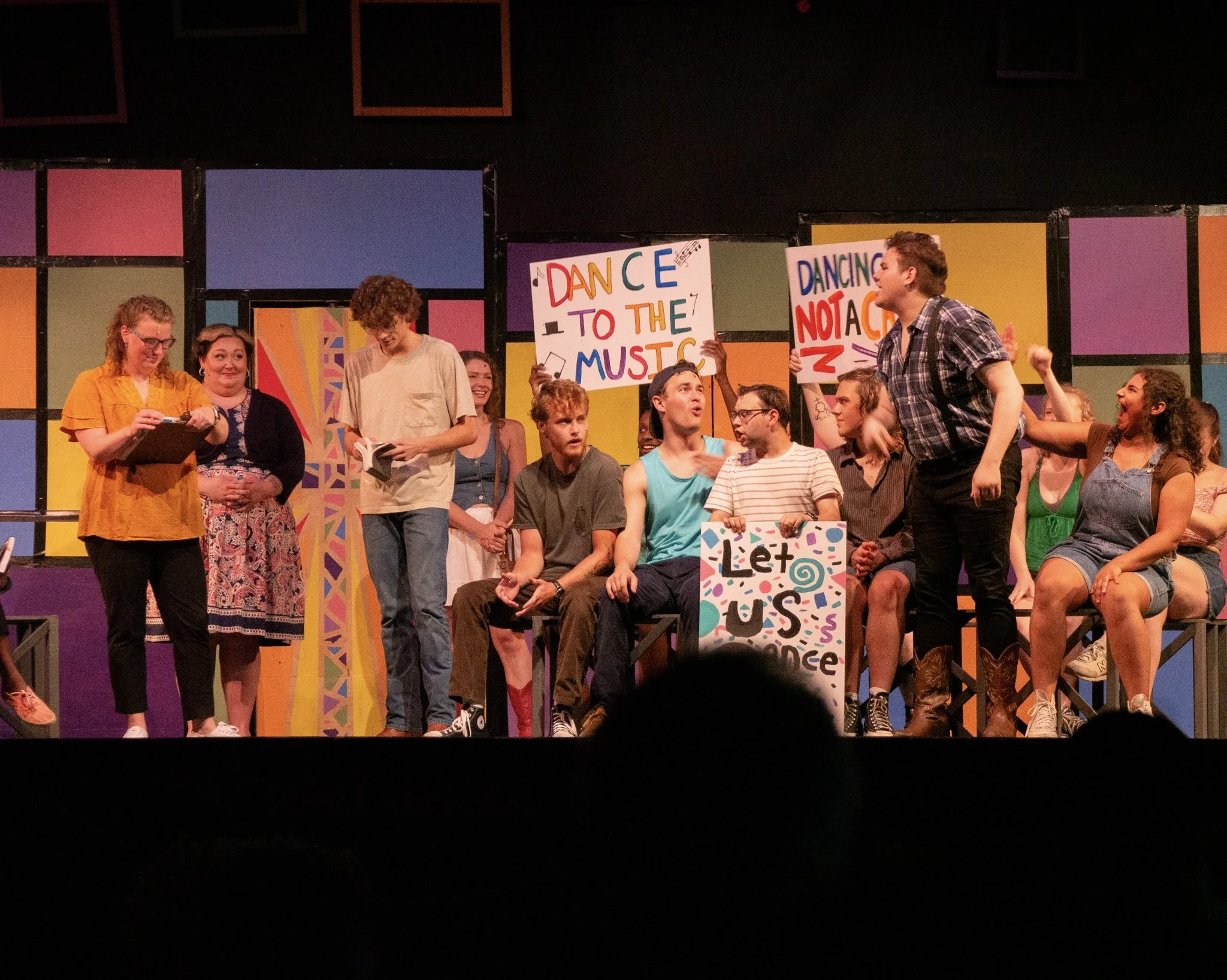 A group of young actors performing on stage in a school play, with colorful geometric backgrounds and signs that say 'Dance to the Music,' 'Dancing Not a Crime,' and 'Let Us Dance.'
