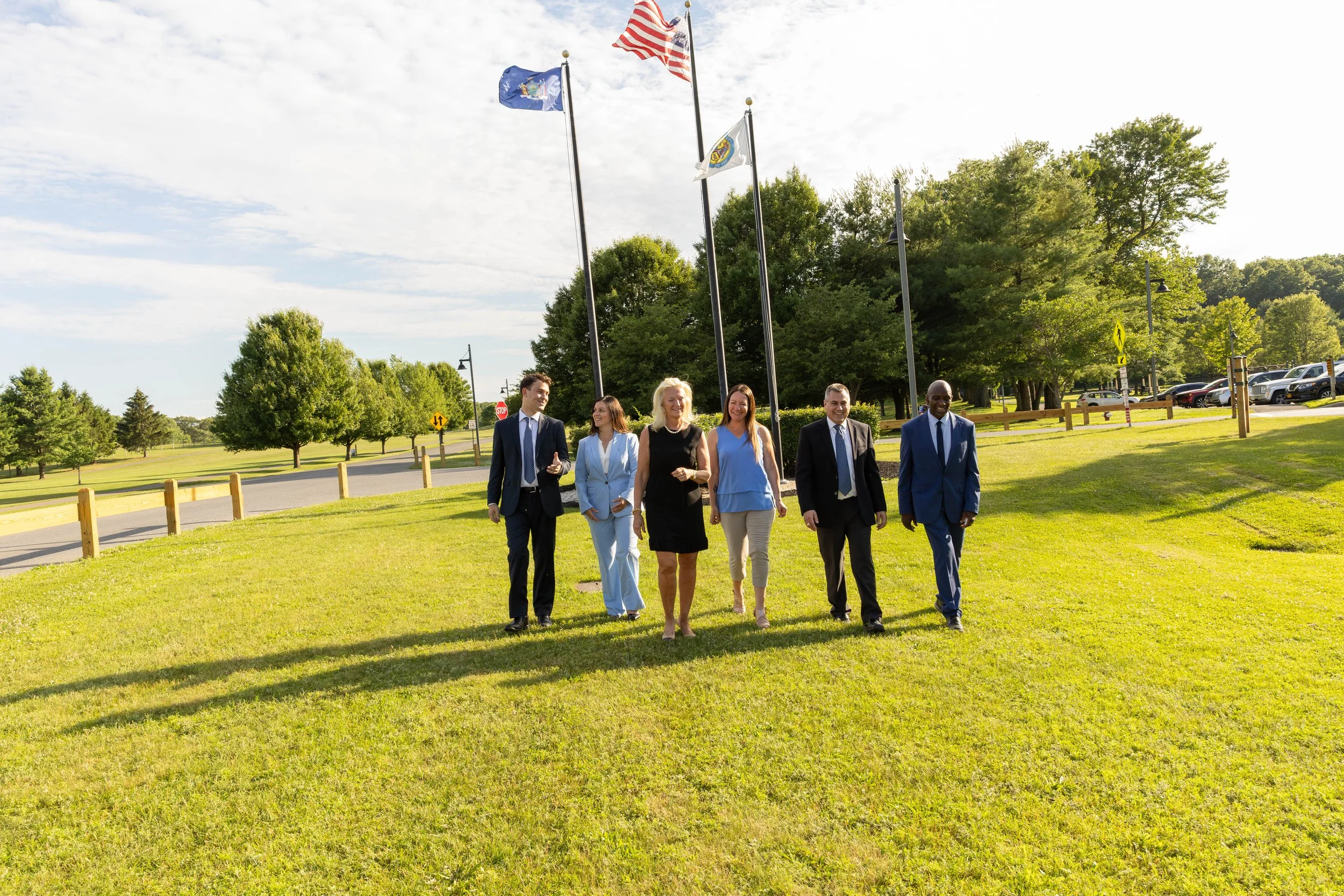 Group of six diverse people, dressed in business attire, walking on a grassy park with flagpoles and trees, on a sunny day.