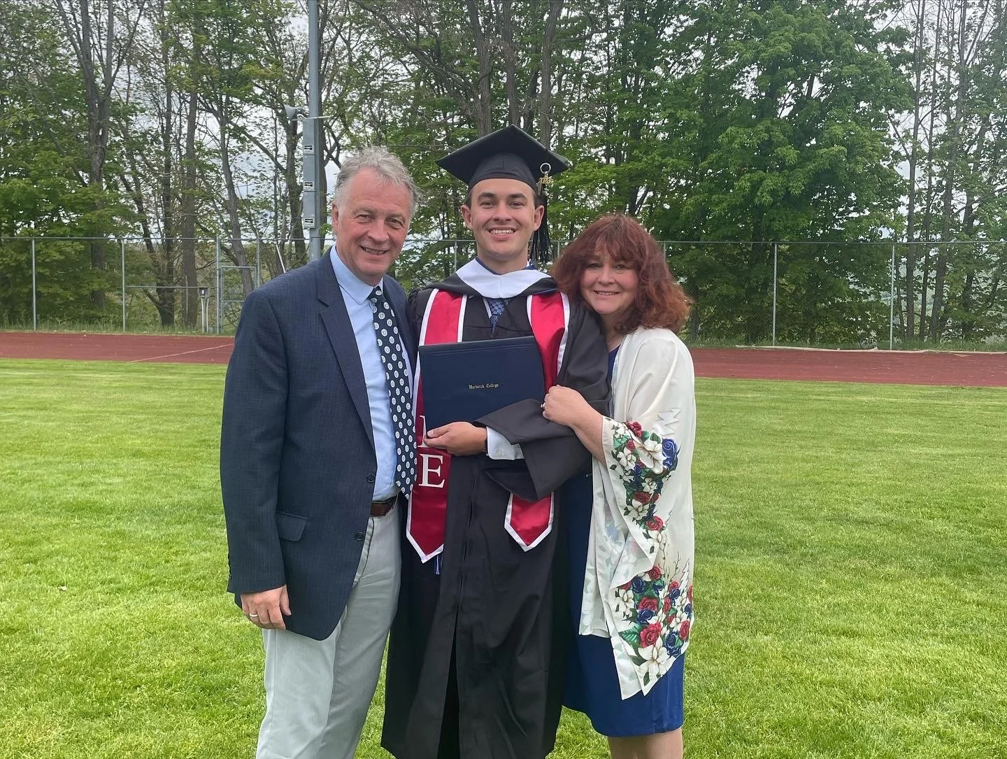A young man in a graduation cap and gown holding a diploma, standing between an older man in a blazer and a woman in a floral shawl, all smiling outdoors on a grassy field.